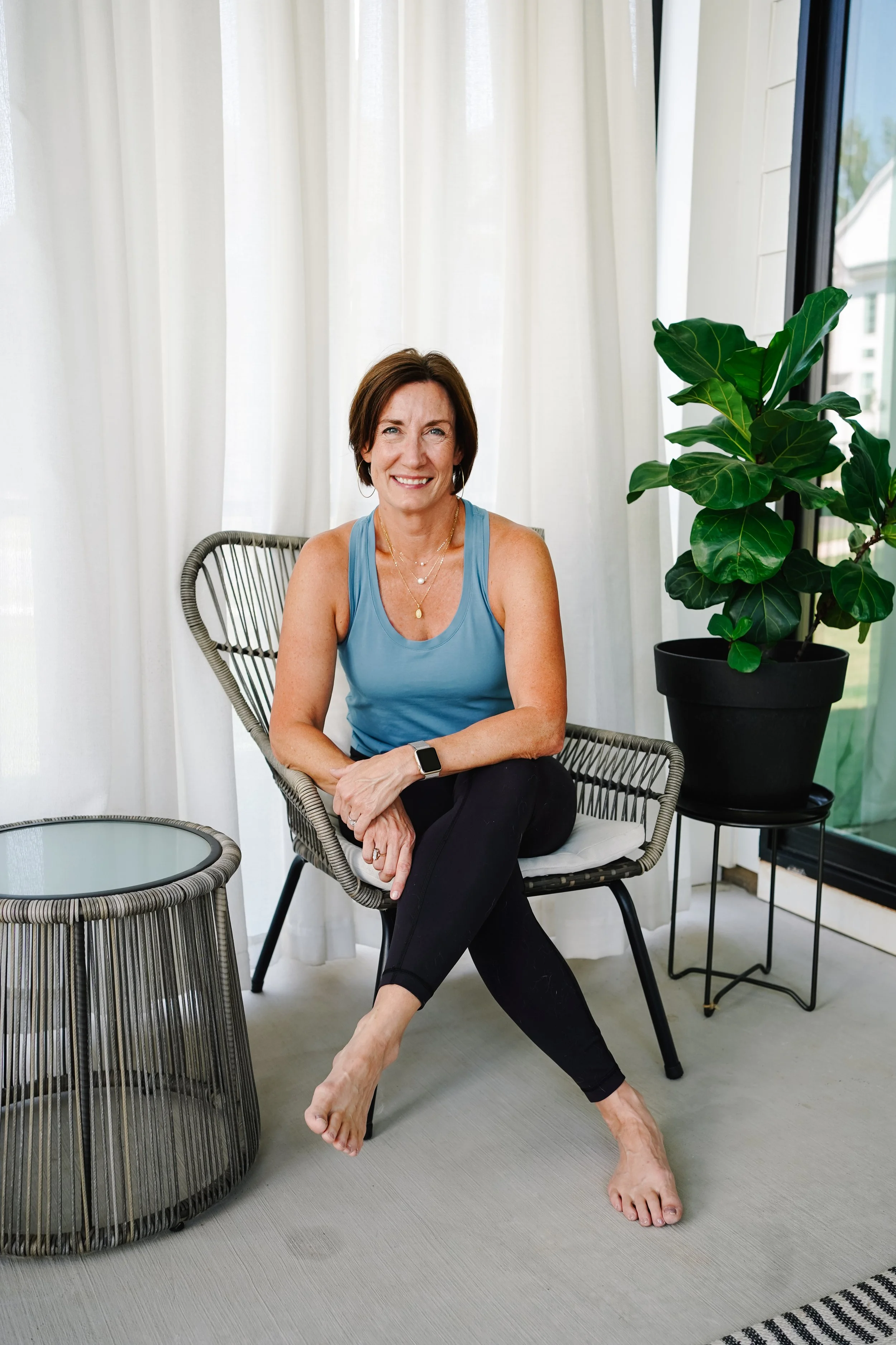 A smiling woman in a blue tank top and black leggings sitting on a modern wicker chair with a white cushion, next to a large potted fig tree on a small black stand, inside a room with white curtains and large windows.