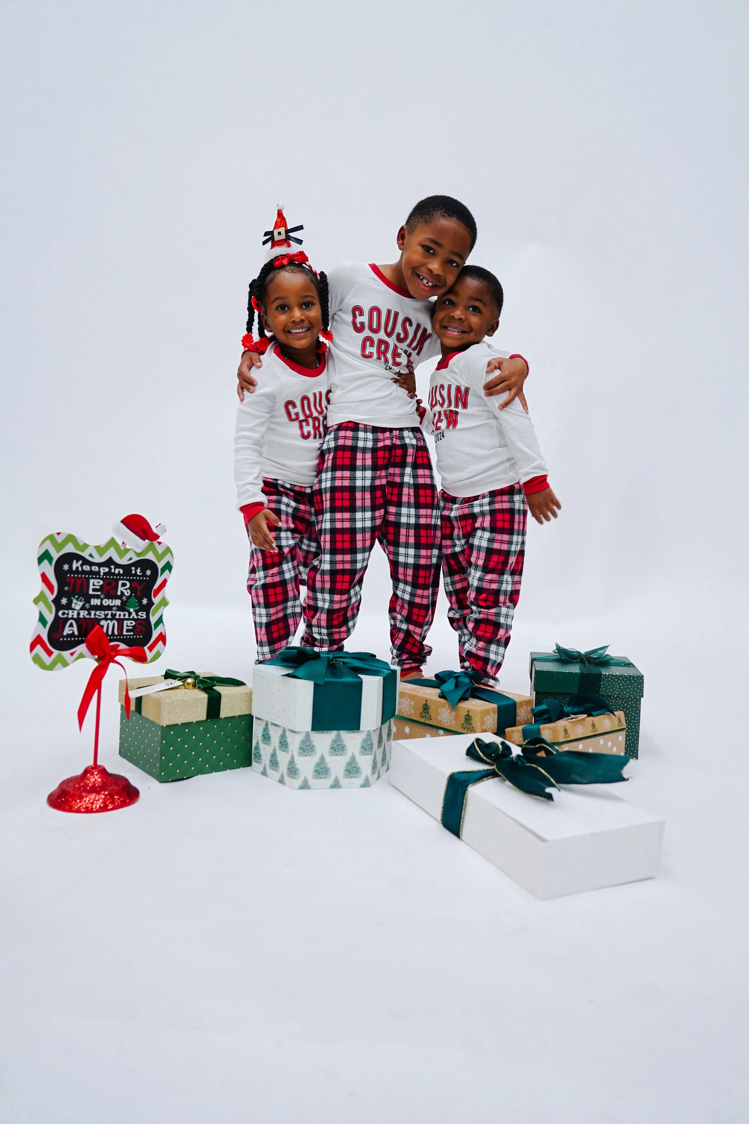 Three children in Christmas pajamas standing on wrapped presents, smiling, with holiday decorations and a sign that says "Keepin it merry in our Christmas jams."