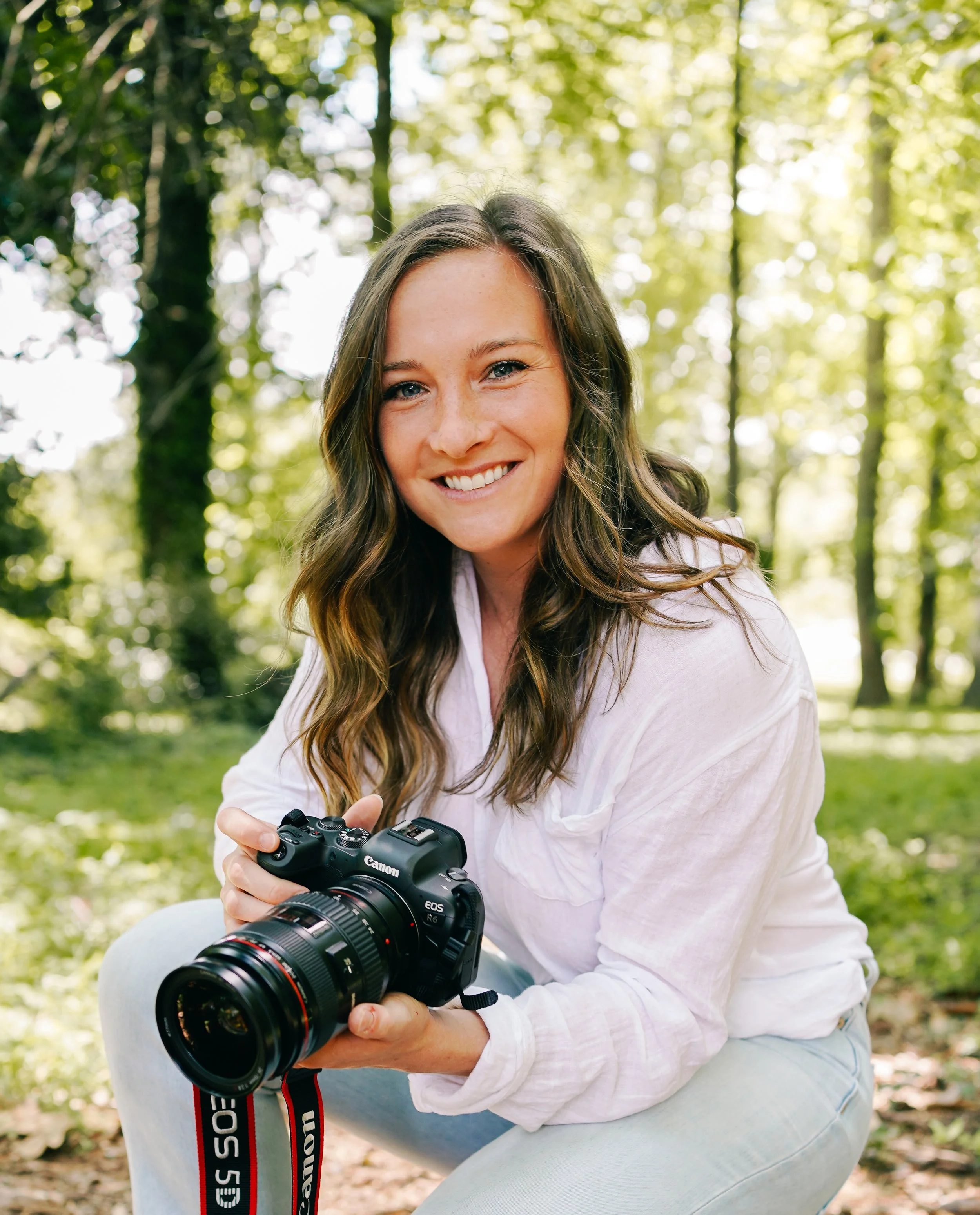 A woman with brown hair and a white shirt holding a Canon camera outdoors in a green, wooded area, smiling at the camera.