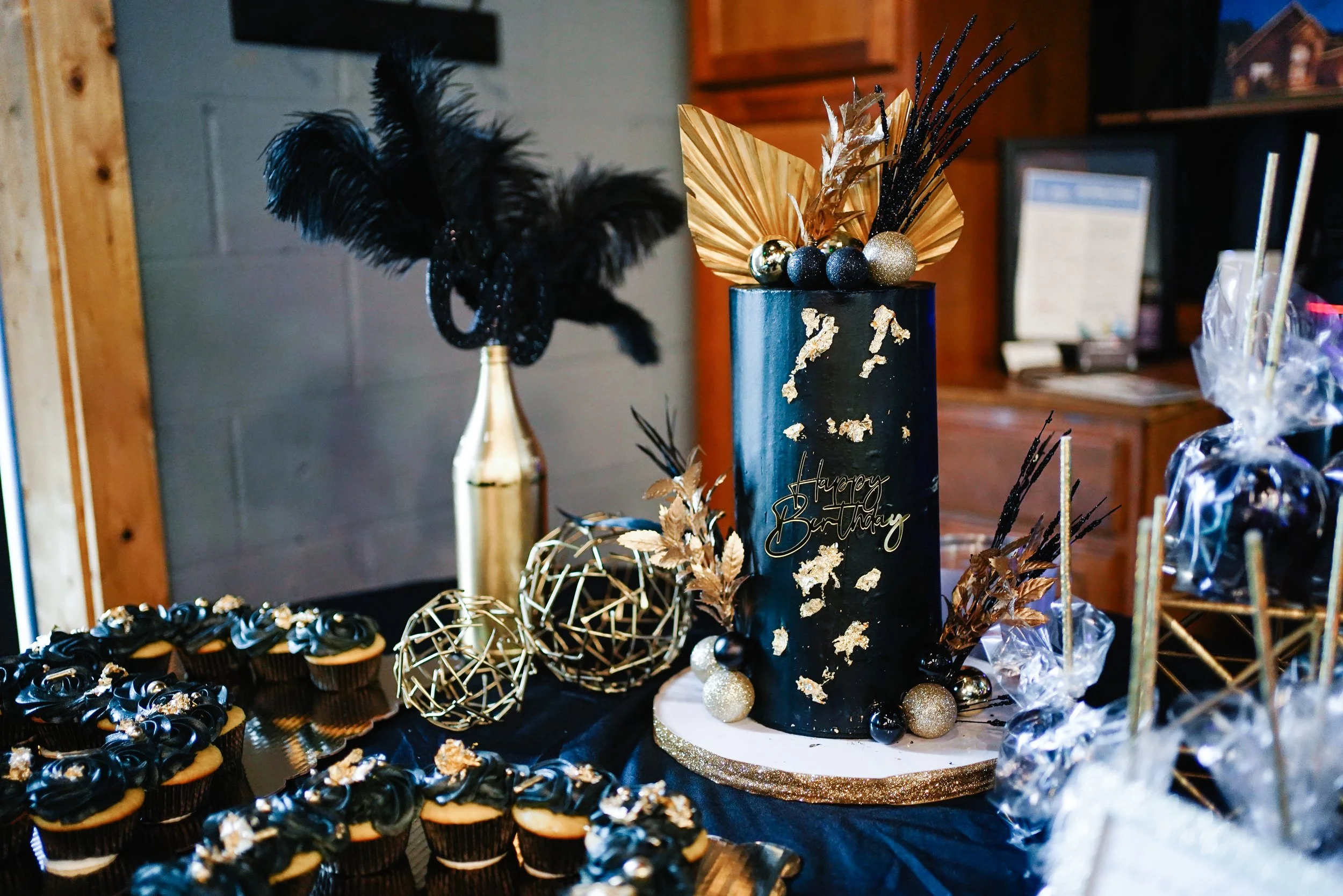 A black birthday cake with gold foil accents and a 'Happy Birthday' message, decorated with gold and black spherical ornaments, golden leaves, and black feathers, surrounded by cupcakes with black frosting and gold embellishments; gold geometric deco