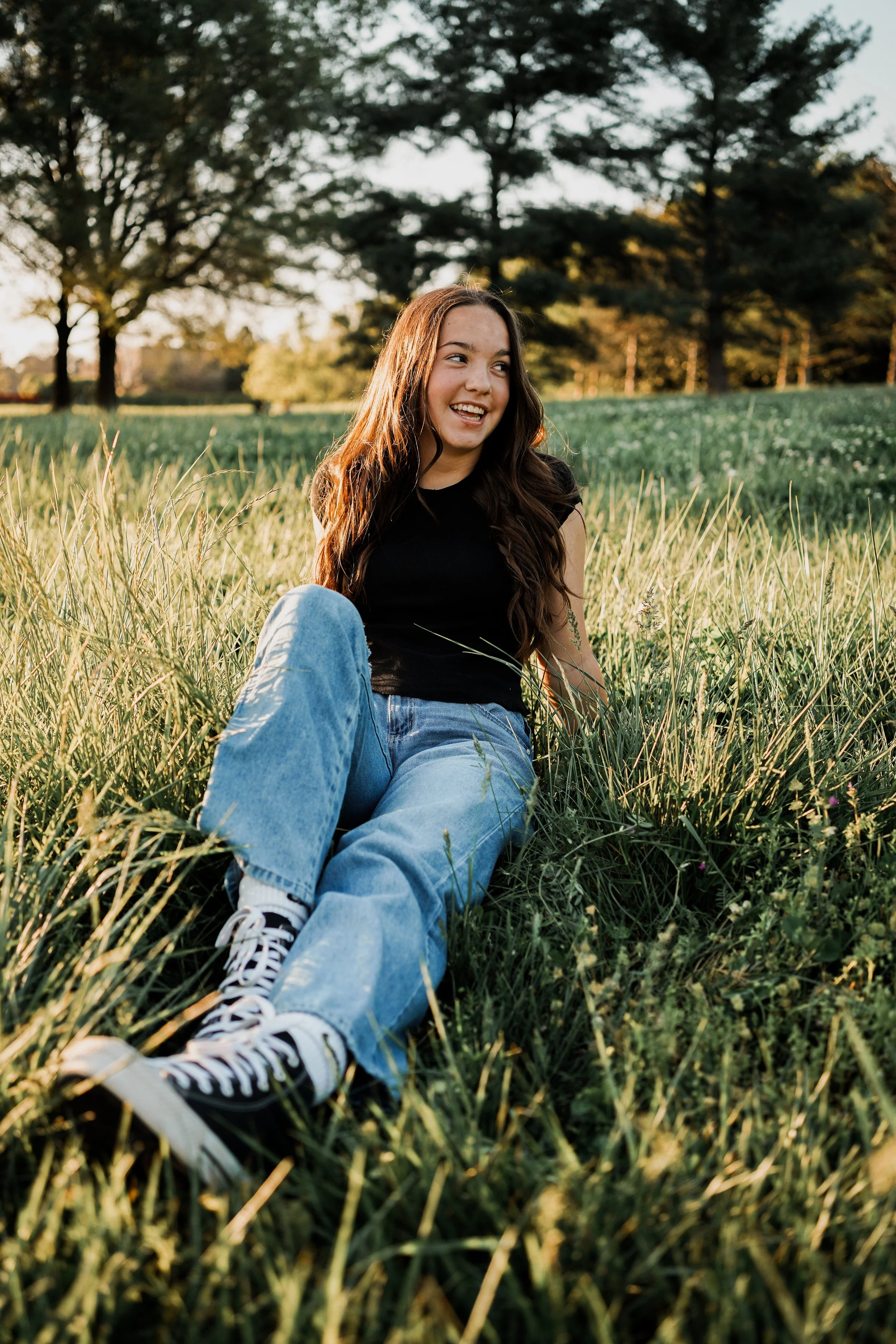 A young woman sitting in tall grass in a park or field during sunset, smiling and looking to the side.