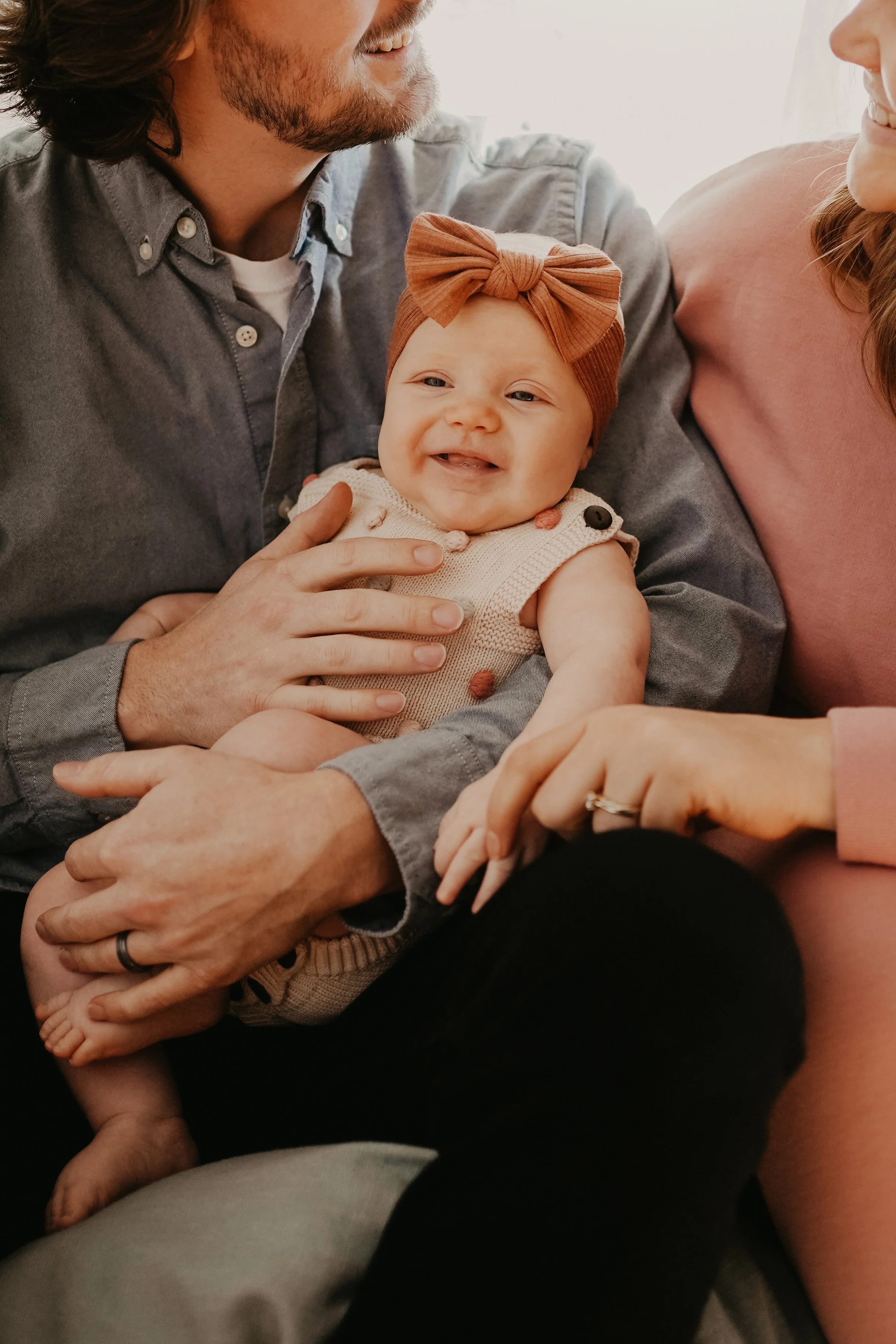 Smiling baby girl wearing a brown headband and sleeveless sweater, being held and cuddled by two adults, likely parents, in a warm, intimate setting.