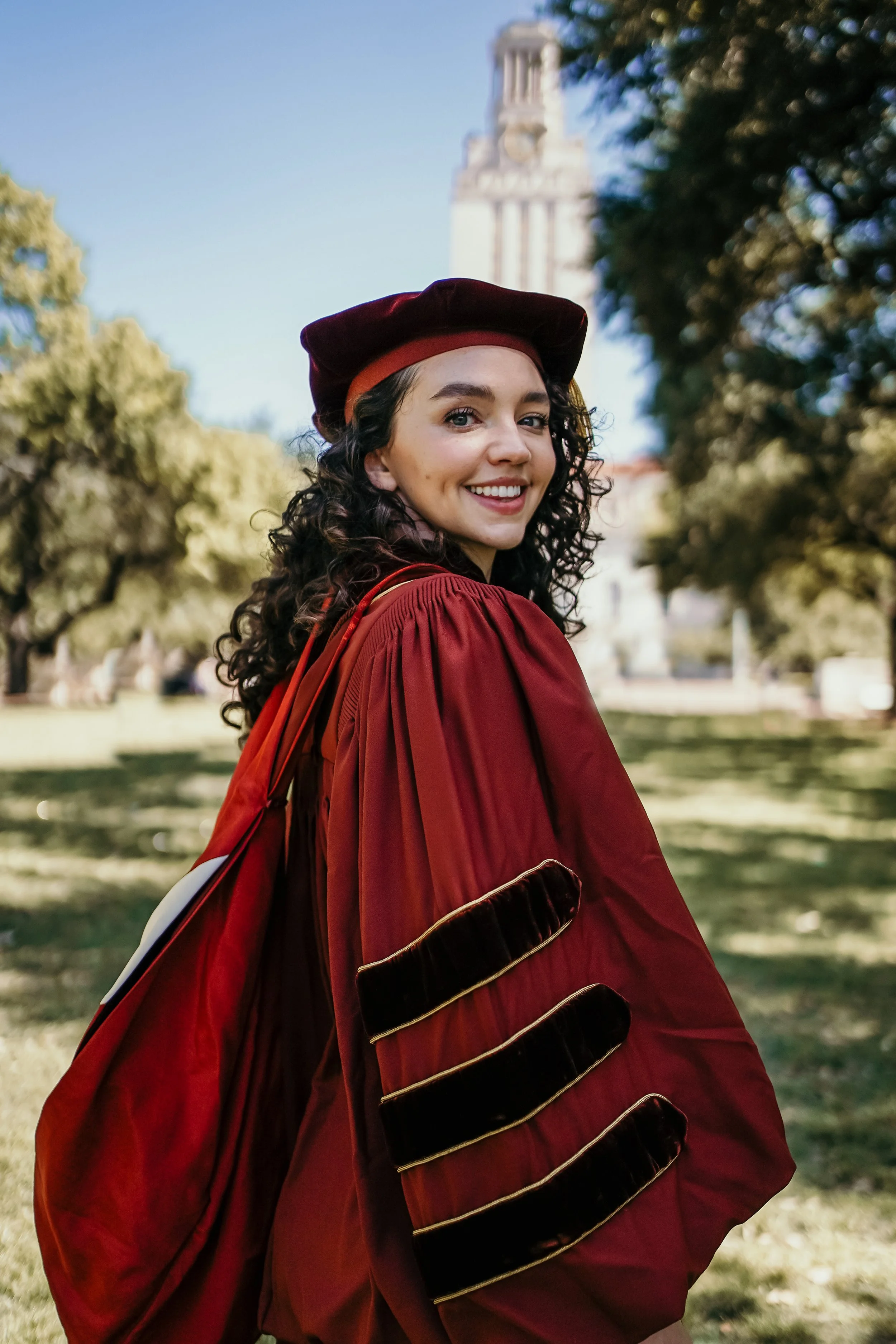 Young woman in a graduation gown and cap, smiling outdoors on a sunny day, with trees and a tall building in the background.