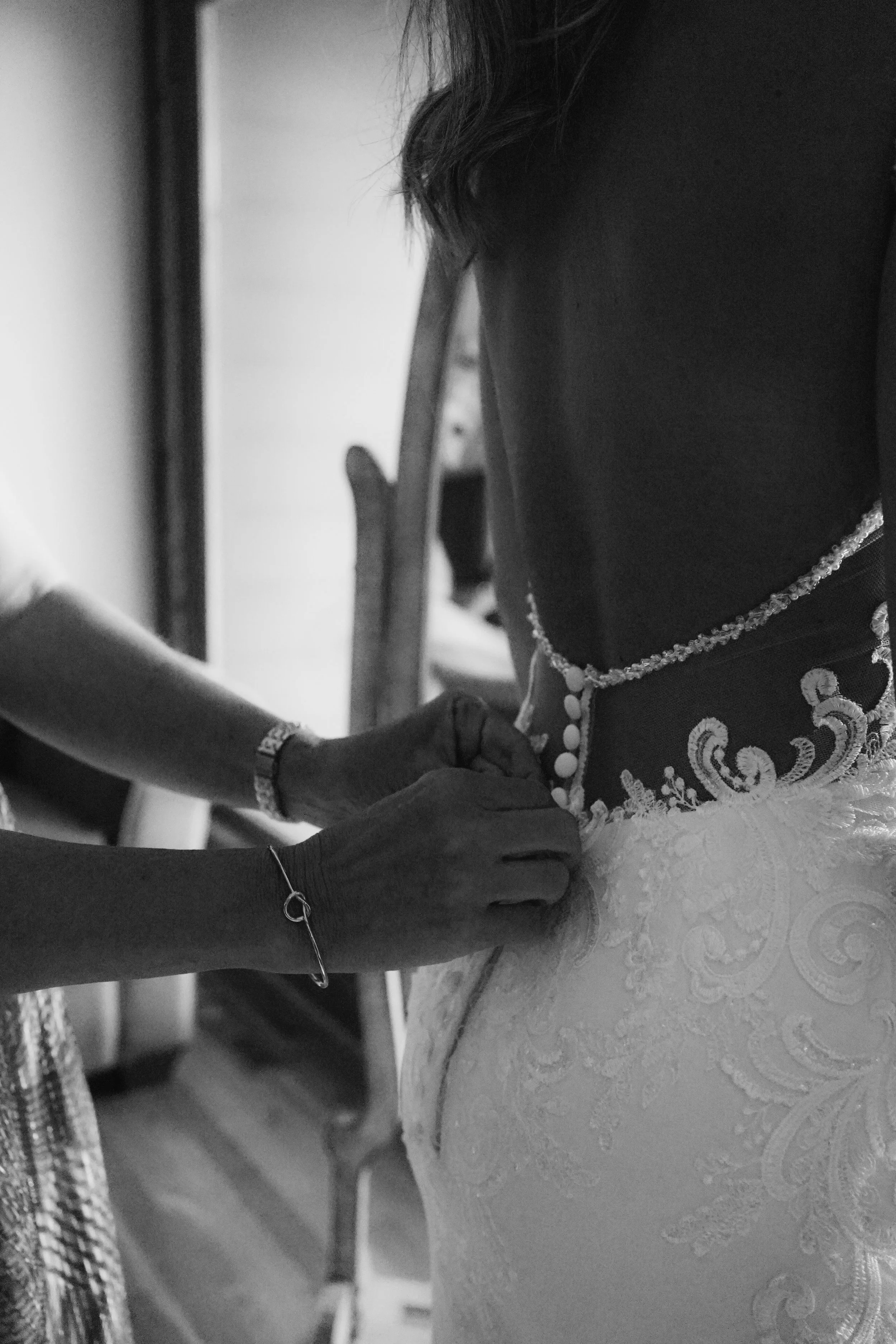 An individual is attaching the back of a wedding dress with buttons while another person assists, in black and white.
