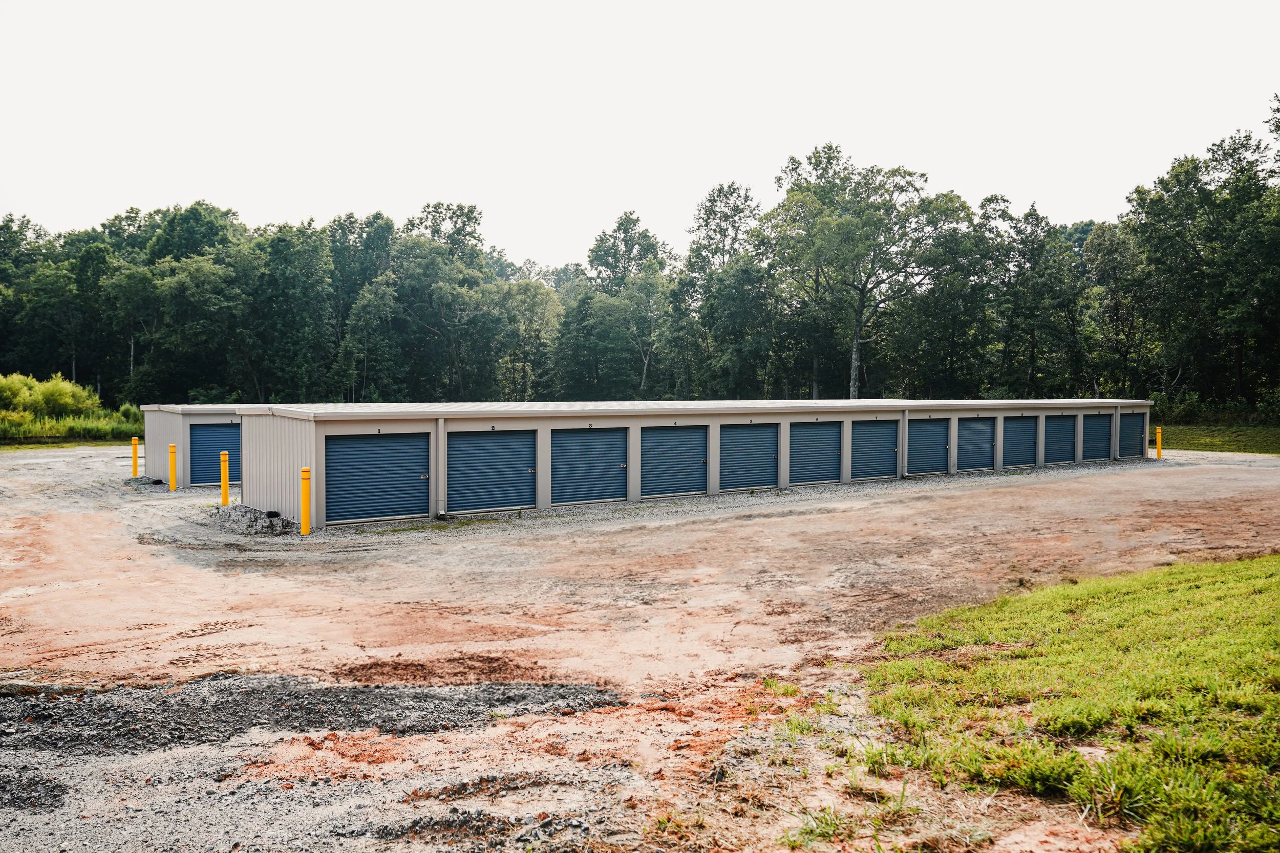A row of storage units with blue roll-up doors in a gravel lot, surrounded by trees.