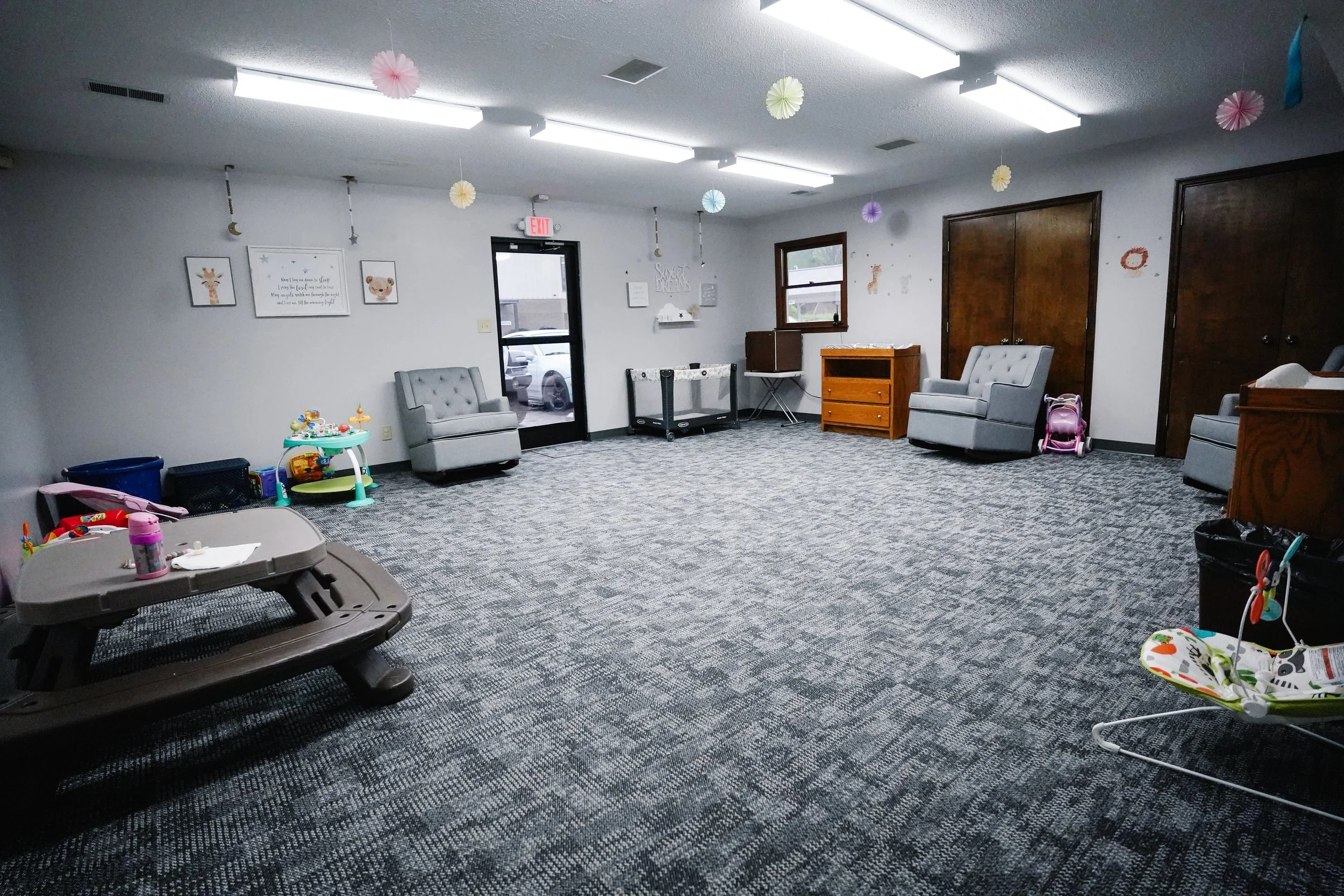 The image shows a spacious room with gray walls and a patterned carpet, decorated for children. There are two gray armchairs, a small wooden dresser, and a play area with toys and a baby swing. The ceiling has colorful paper decorations hanging from 