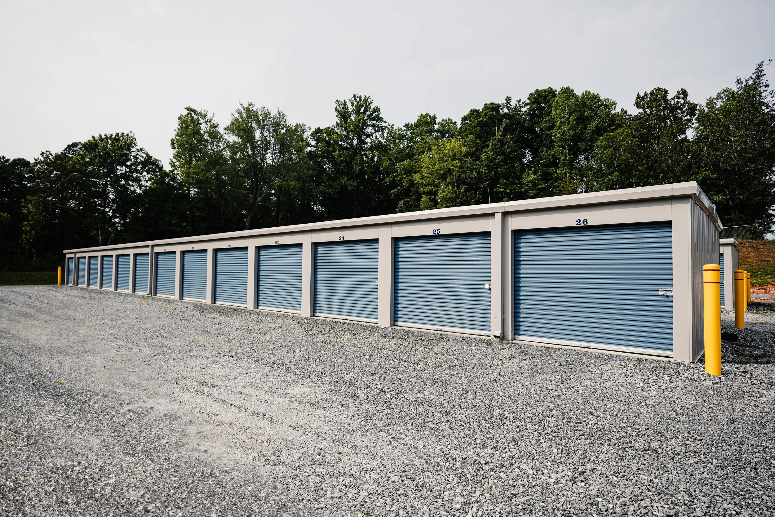 A row of blue storage unit roll-up doors with yellow protective posts on a gravel lot, numbered 21 to 26, with trees in the background.