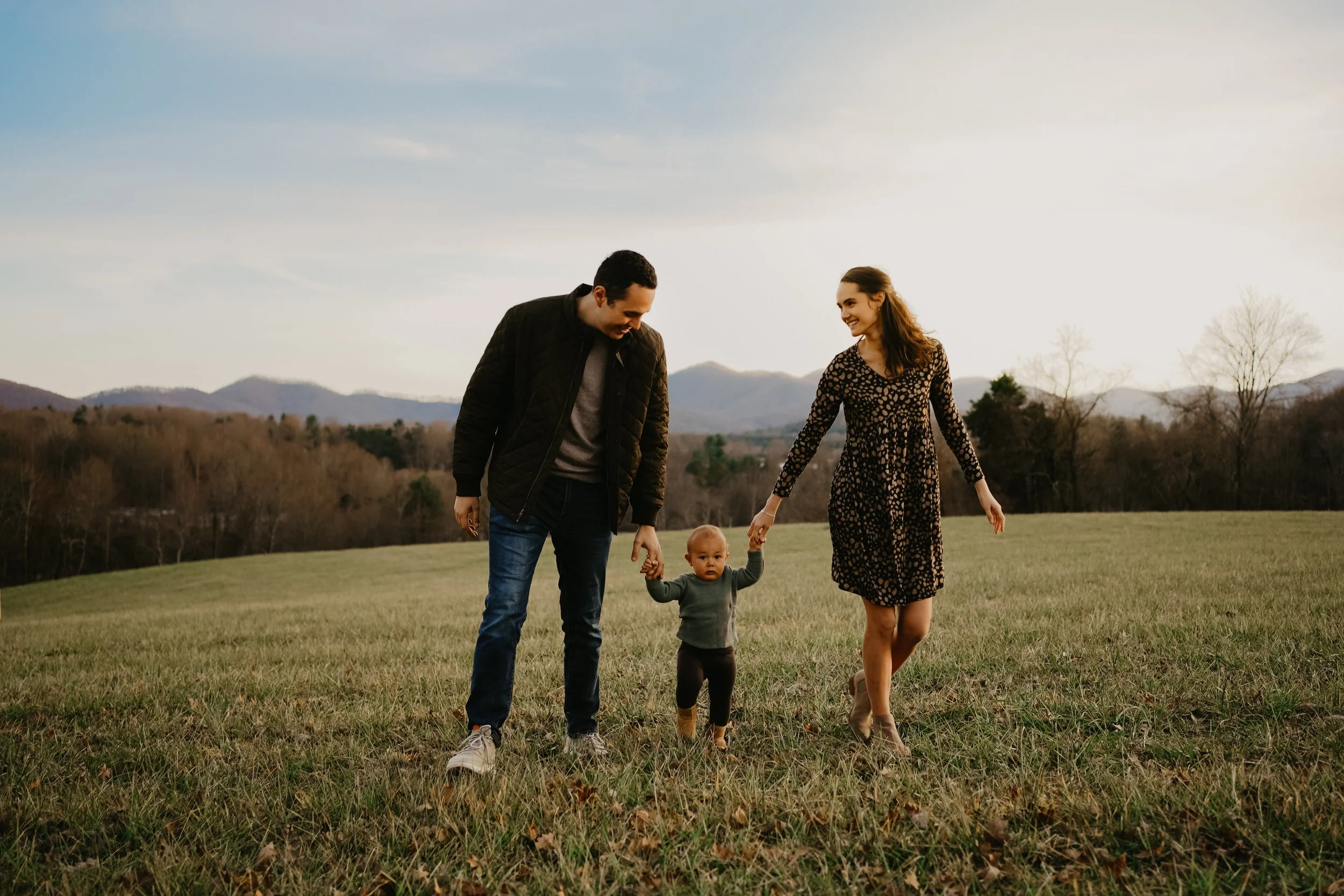 A family of three, a man, woman, and a toddler, walking hand in hand across a grassy field with mountains in the background during late afternoon or early evening.