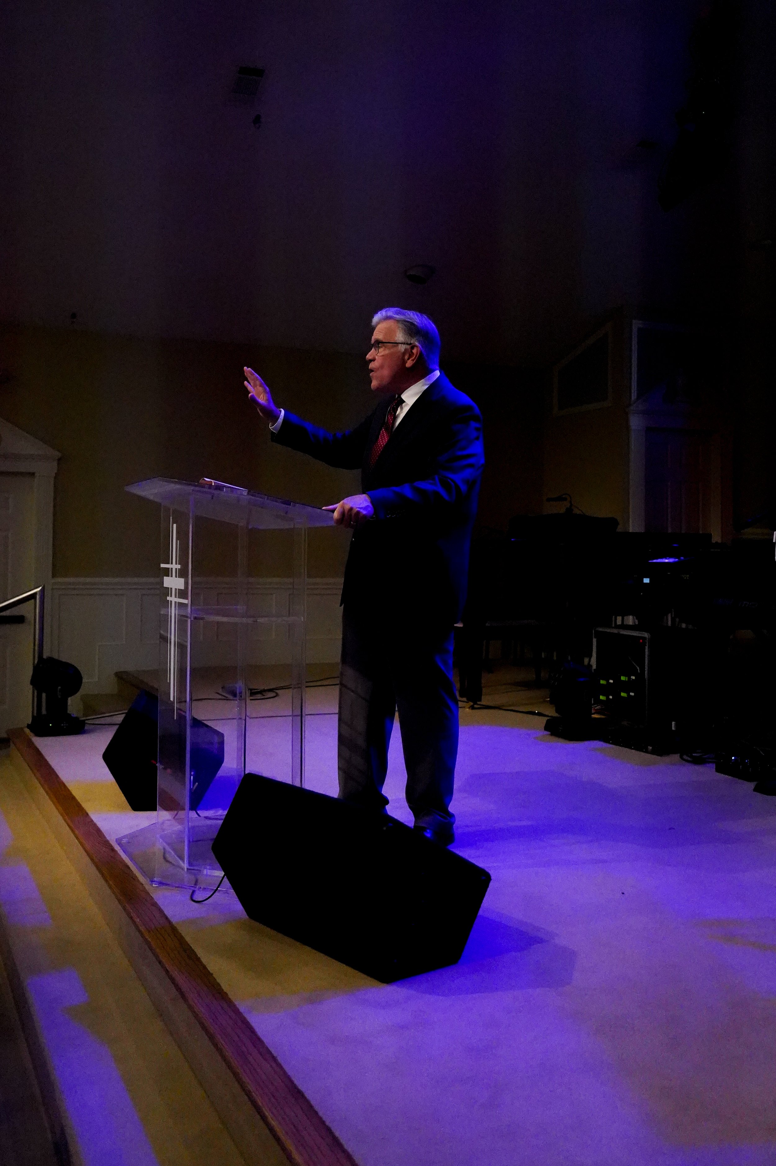 Man in dark suit standing on stage behind clear podium, speaking or giving a presentation with his hand raised, in a dimly lit room with audio equipment and a black speaker in front.
