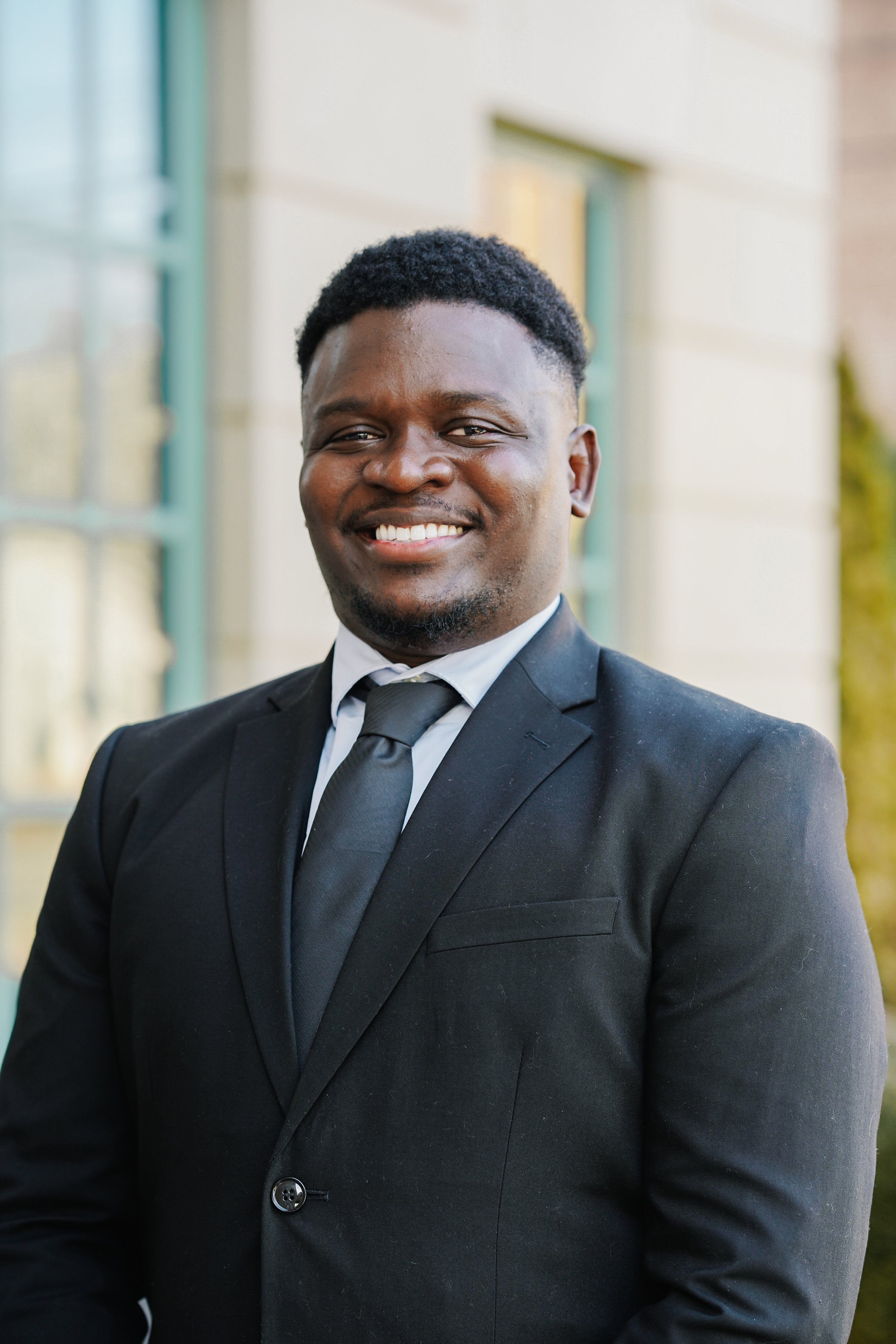 A smiling man in a black suit, white shirt, and black tie posing outdoors in front of a building with large windows.