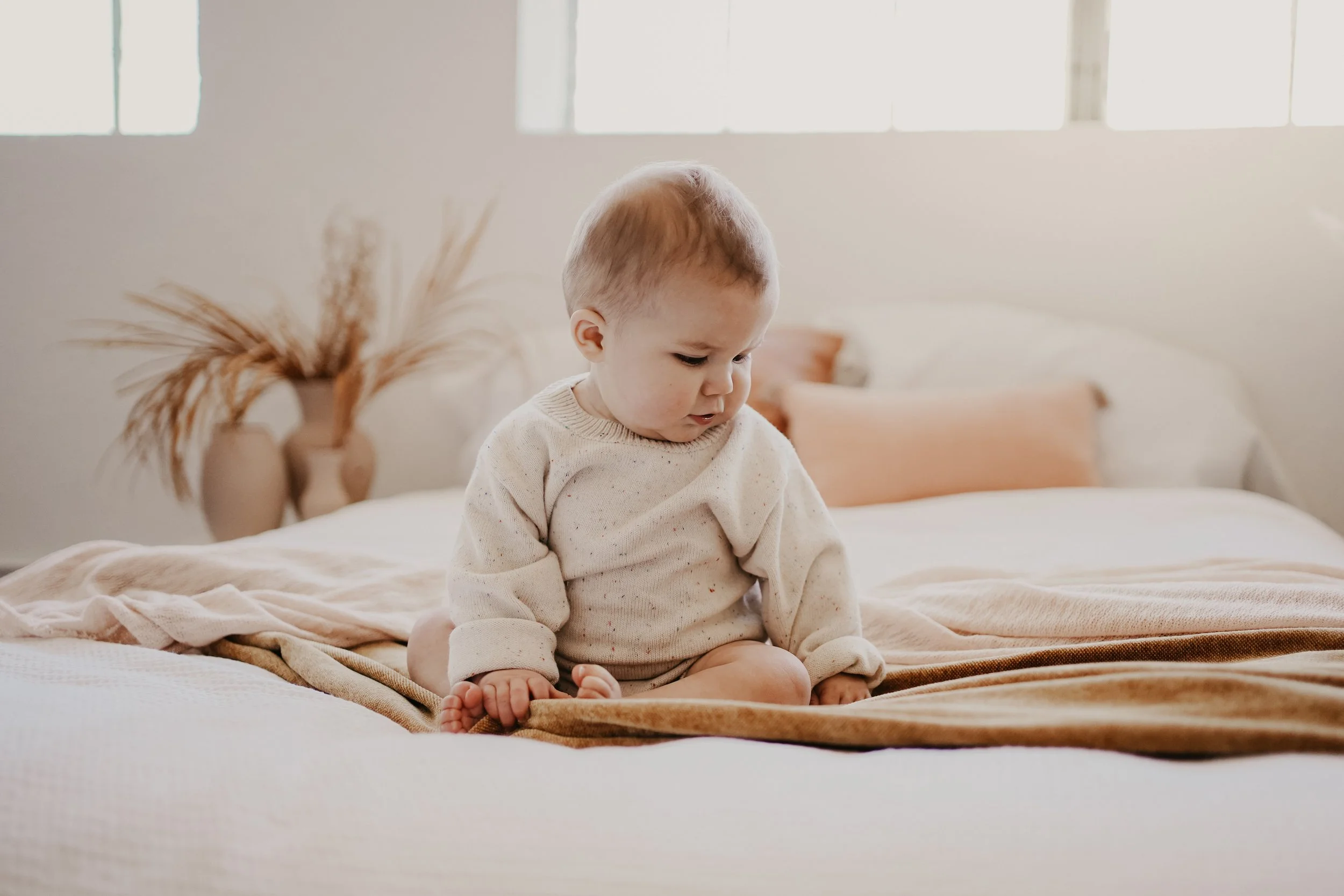 A young child sitting on a bed in a sunlit bedroom, wearing a beige sweater, with a neutral expression.