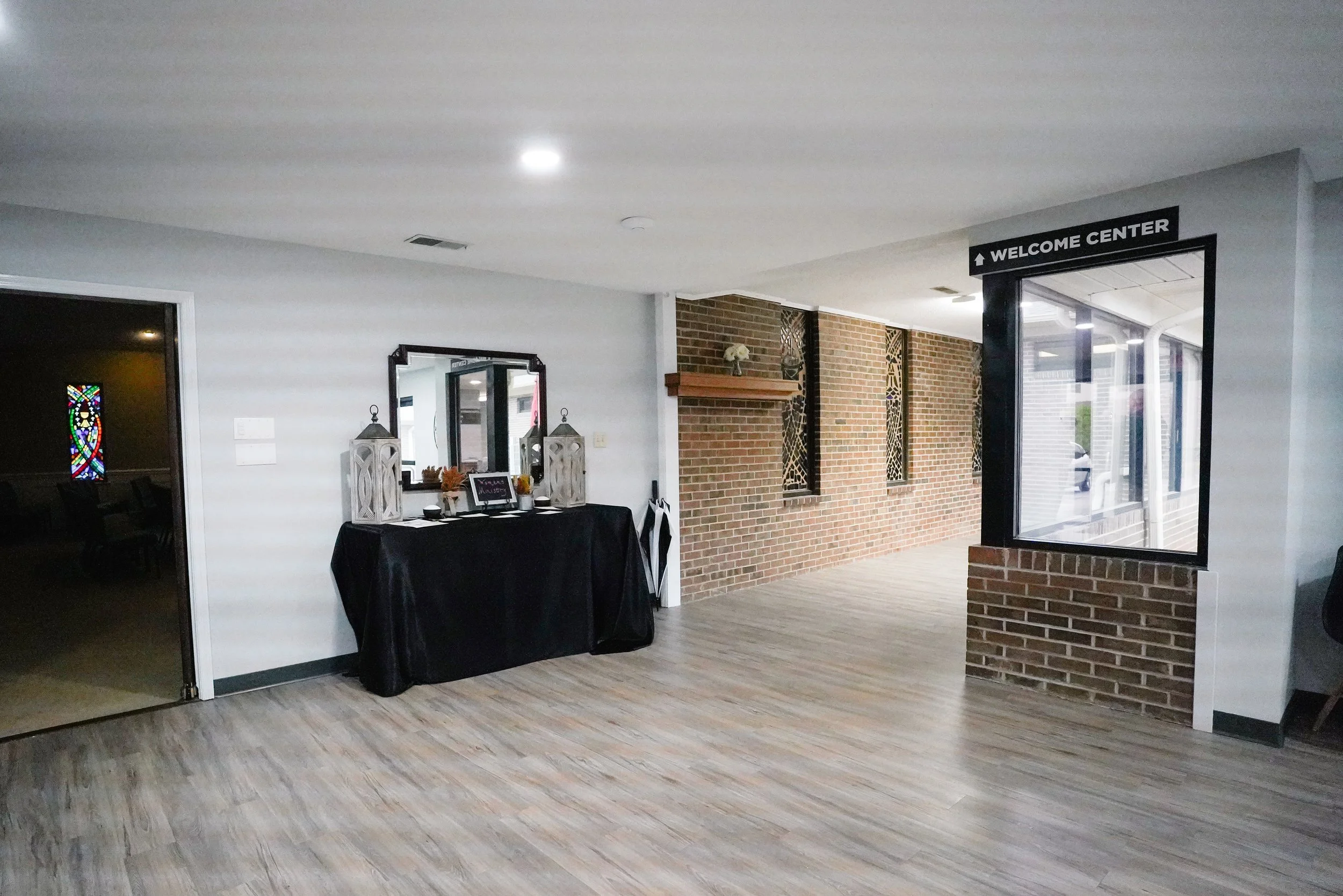 Interior of a welcoming lobby with a table decorated with lanterns and flowers, a mirror, a brick wall with a wooden shelf and decorative panels, and a large window with a sign that reads 'Welcome Center'.