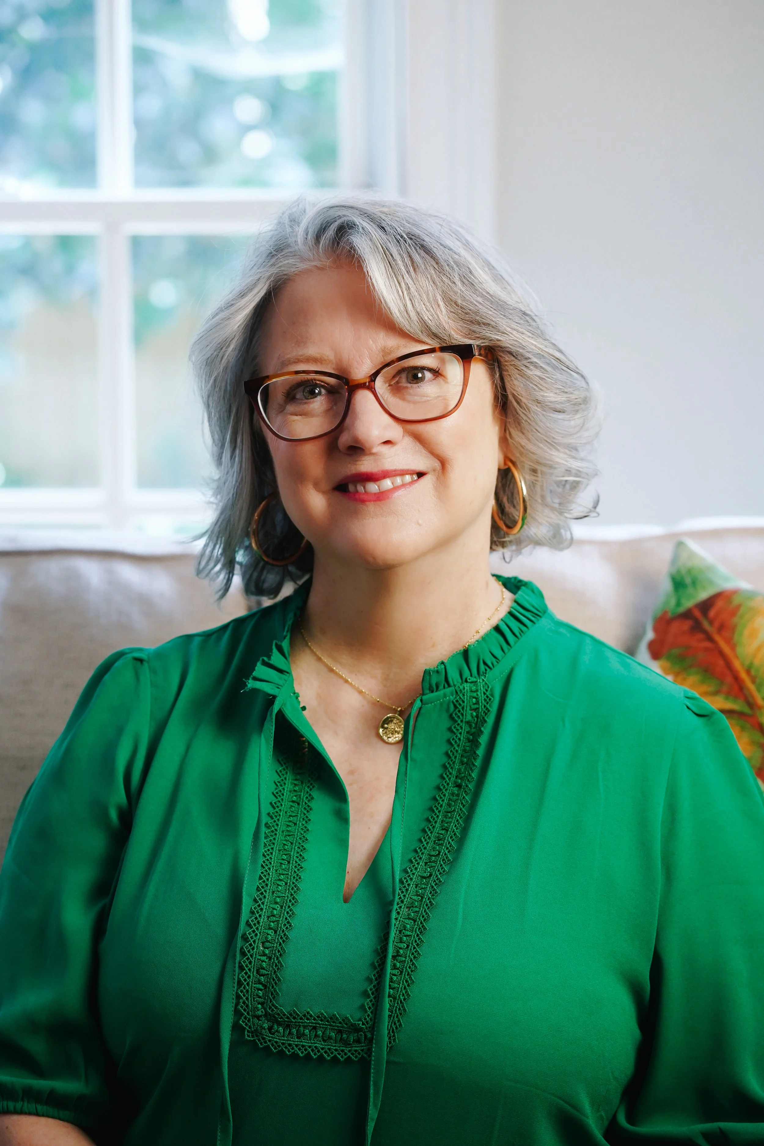 A woman with gray hair wearing glasses, gold jewelry, and a green blouse, sitting on a beige sofa with a colorful pillow, in front of a window with a blurred outdoor background.