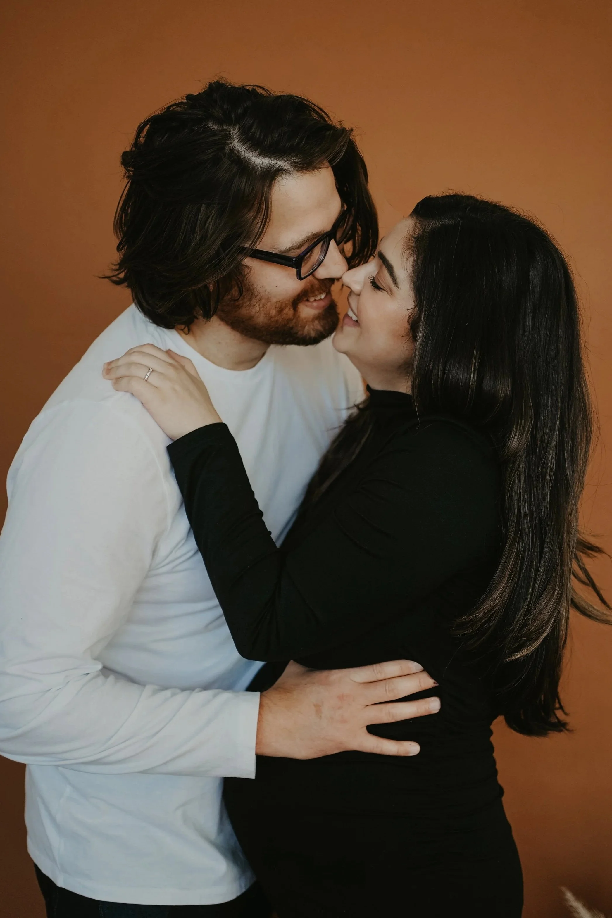 A man and a woman are close together, smiling and about to kiss. The man has glasses and long dark hair, and the woman has long dark hair and is wearing a black outfit. They are embracing each other against a plain brown background.