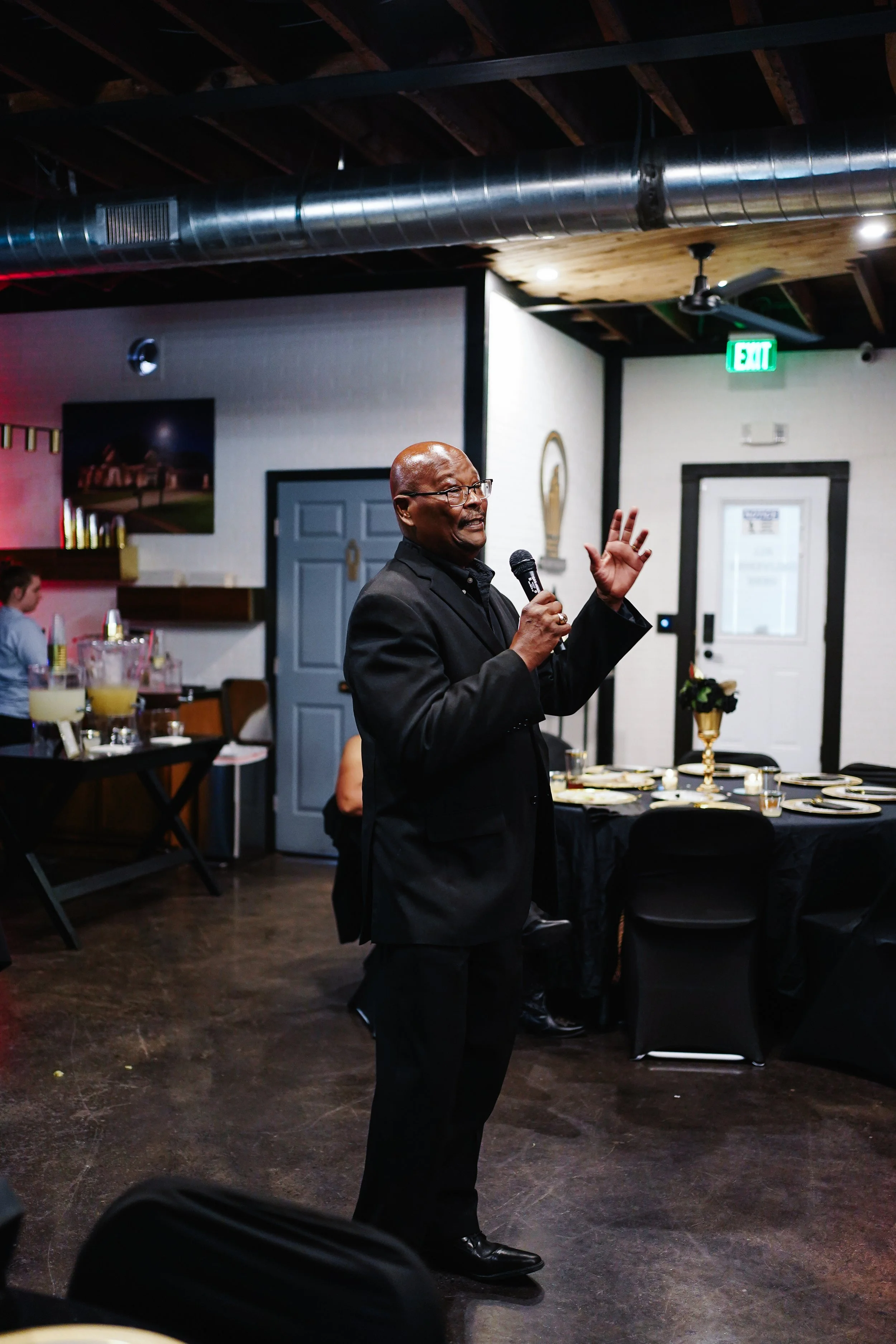 A man in a black suit speaking into a microphone during an indoor event, with a decorated table set for a meal behind him.