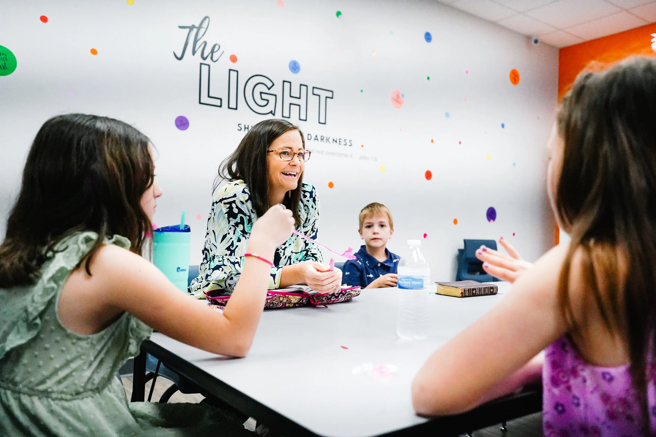 A woman leading a children's Bible study session at a table with three kids, against a wall with 'The Light' written on it, colorful dots, and a Bible resting on the table.