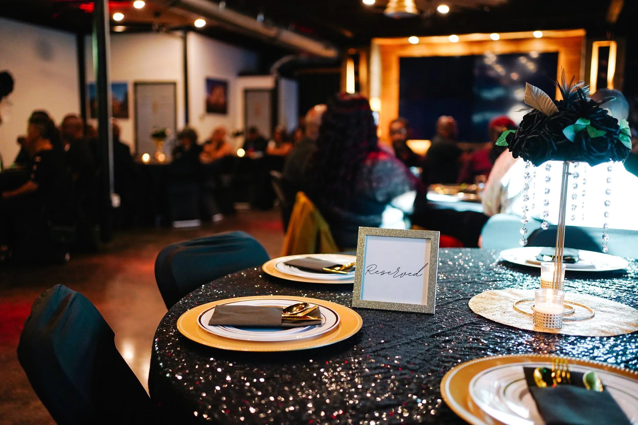 Celebration table decorated with black sequined tablecloth, gold and white dinnerware, and a floral centerpiece with black flowers, feathers, and hanging beads, in a dimly lit event space with many guests.