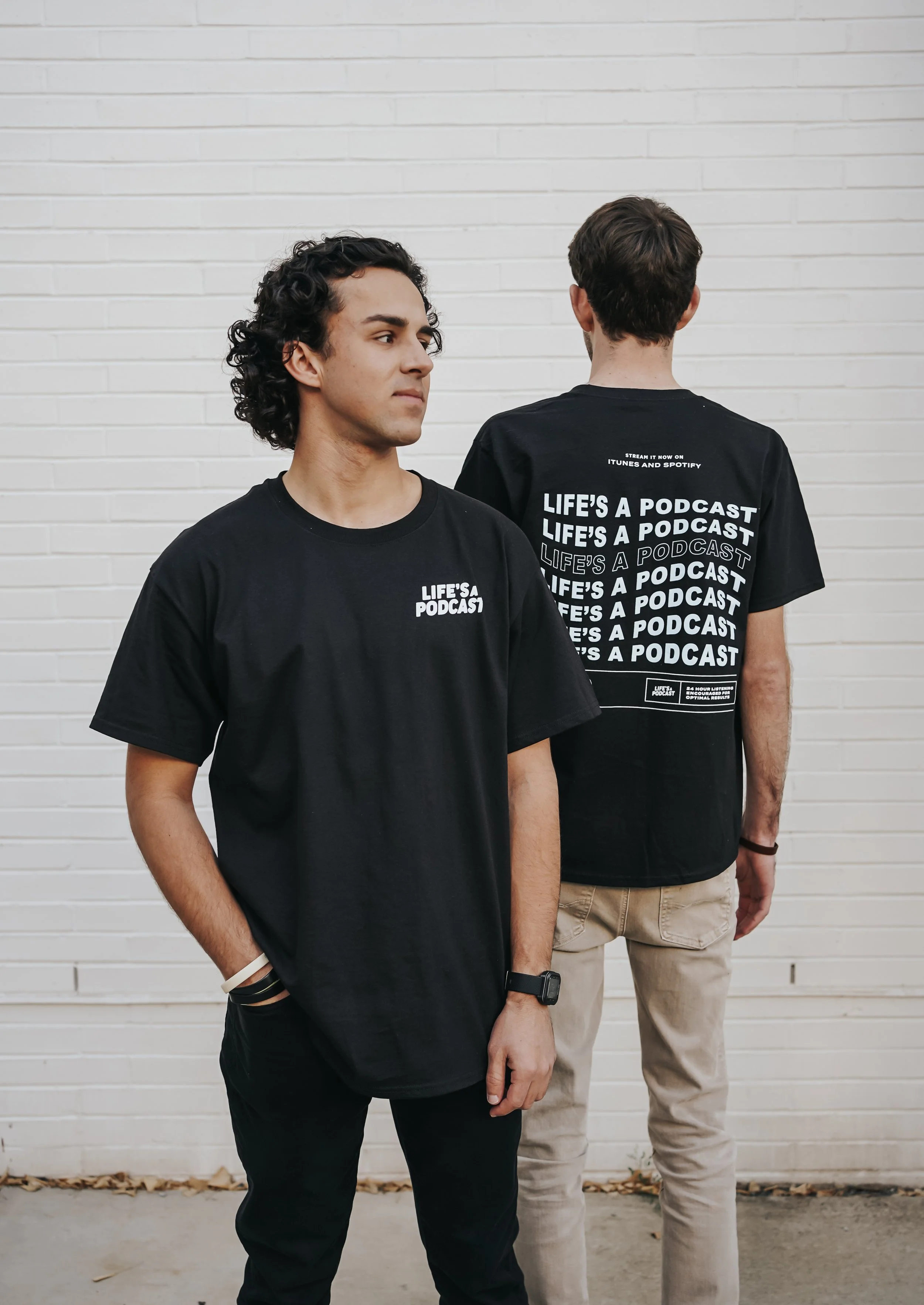 Two young men standing against a white brick wall, wearing black t-shirts with text related to a podcast, one looking to the side and the other facing away.