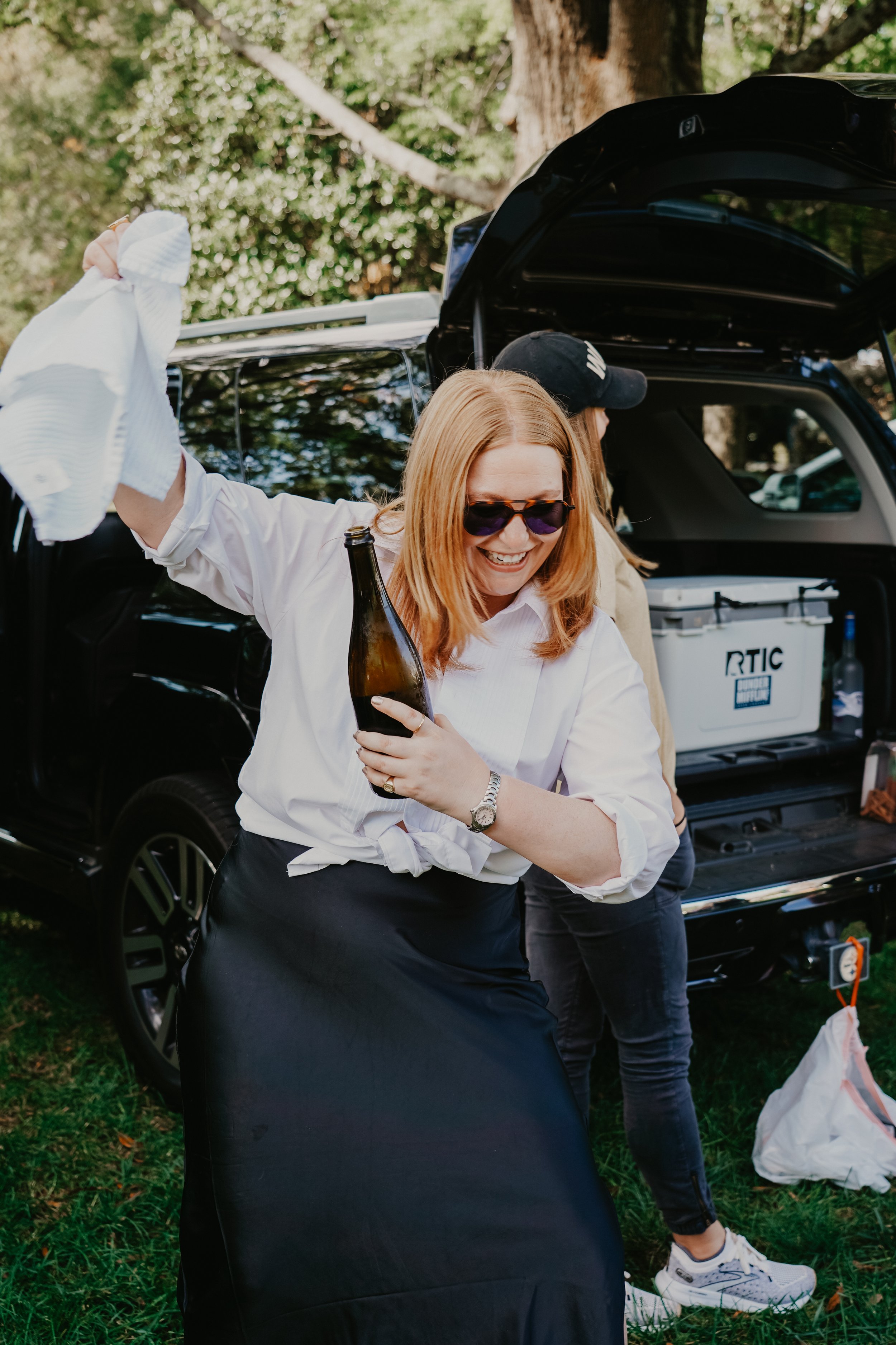 A woman with red hair, sunglasses, a white shirt, and a black skirt is smiling and holding a beer bottle and a cloth, dancing outdoors near a black car with an open trunk, with a person in a beige jacket and black cap partially visible behind her. Th