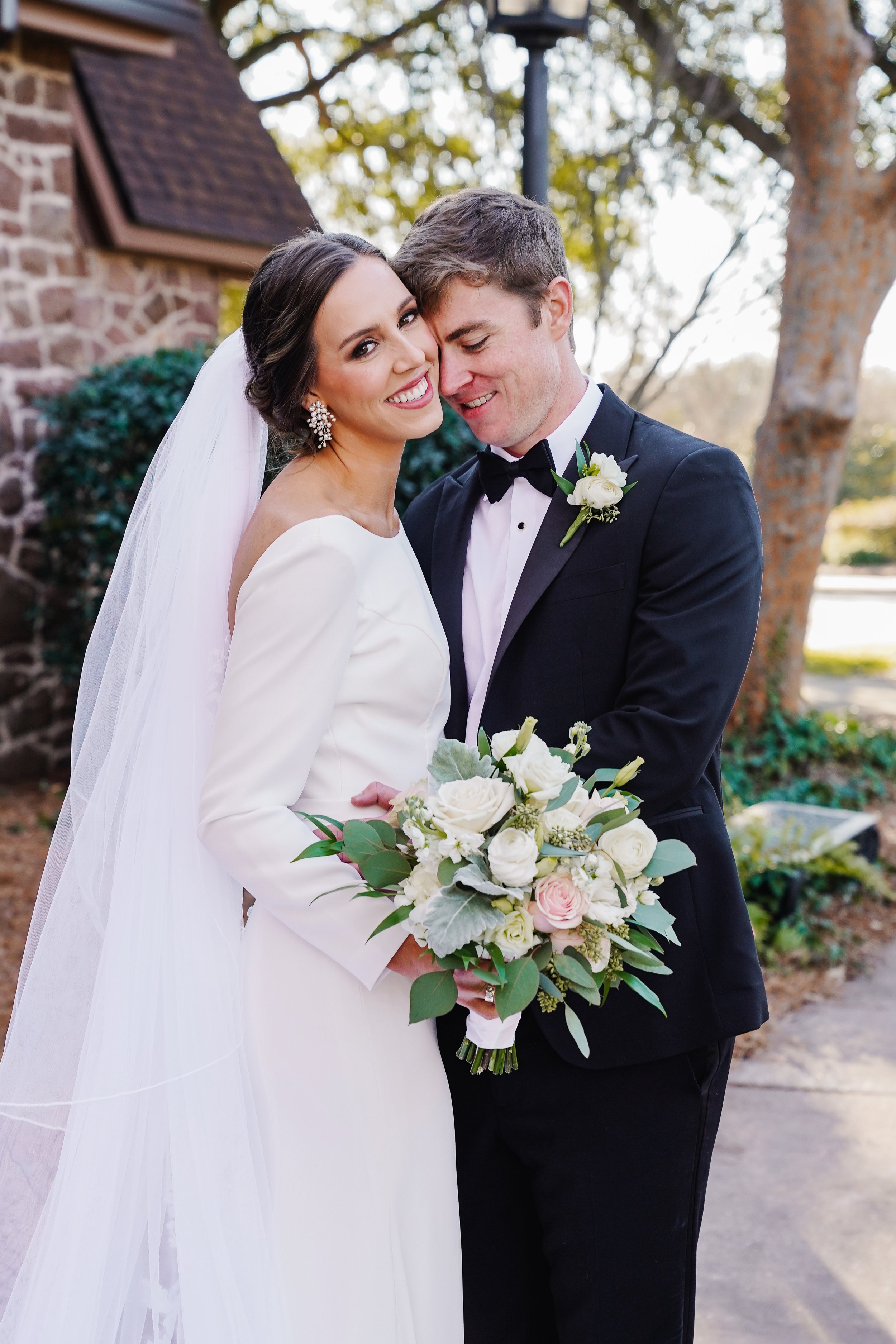 Bride and groom embrace outdoors, smiling, with the bride holding a white and pink floral bouquet, dressed in white, and the groom in a black tuxedo with a white boutonniere.