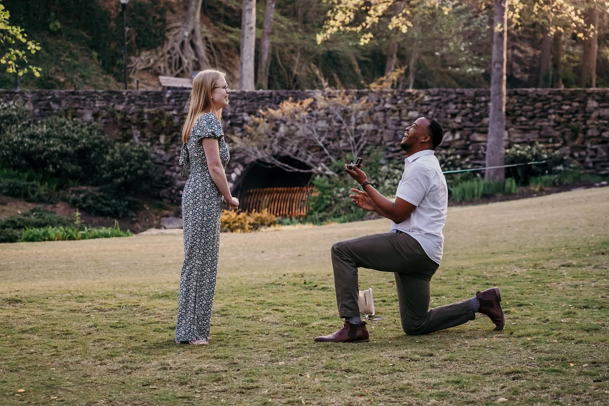 A man down on one knee proposing to a woman in a park with trees and a stone wall in the background.