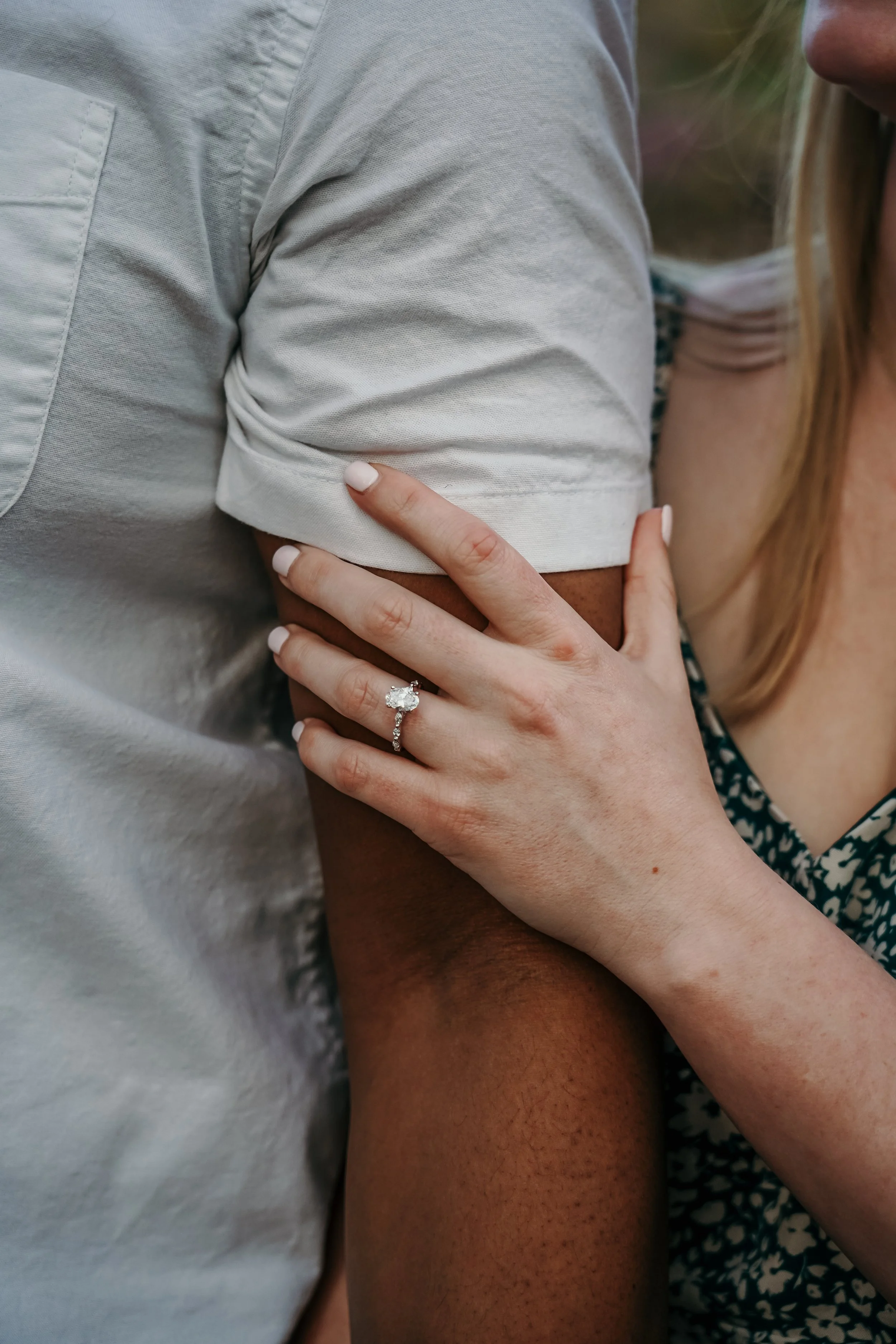 Close-up of a woman’s hand with a diamond engagement ring resting on a person’s arm during an outdoor moment.