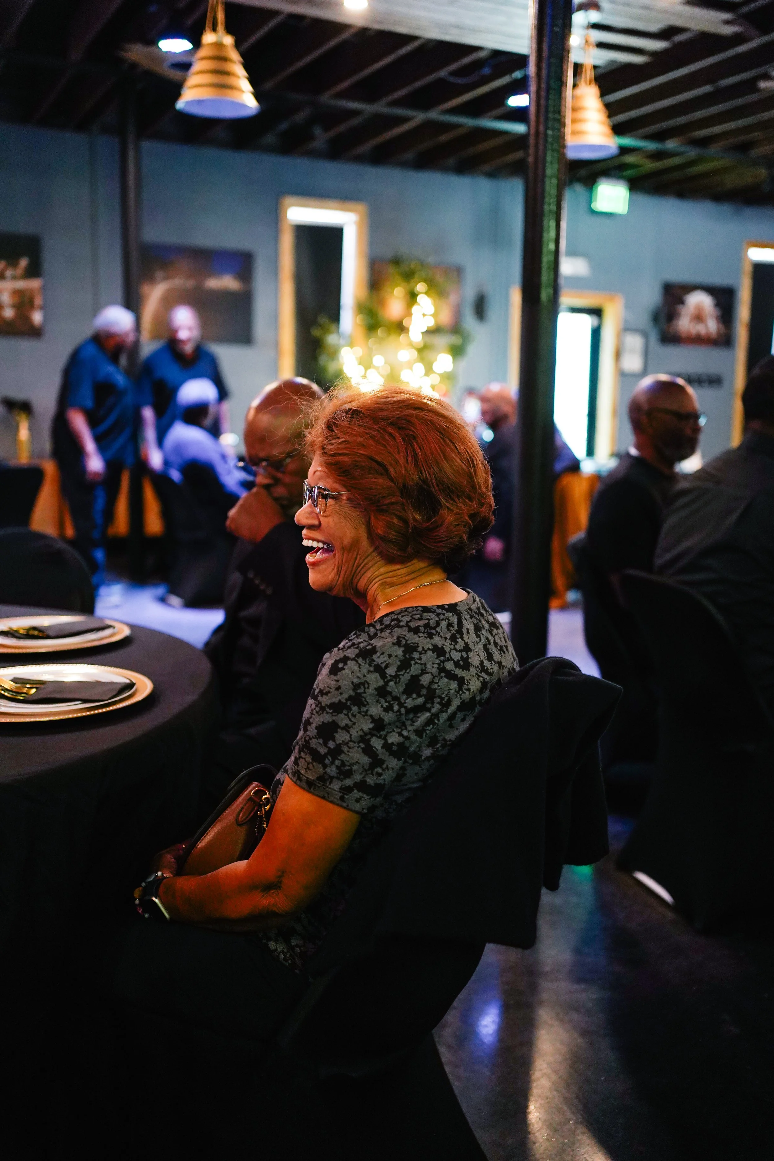 A woman with short ginger hair and glasses, smiling during a gathering or celebration at an indoor venue with dark walls, decorated with lights and artwork.