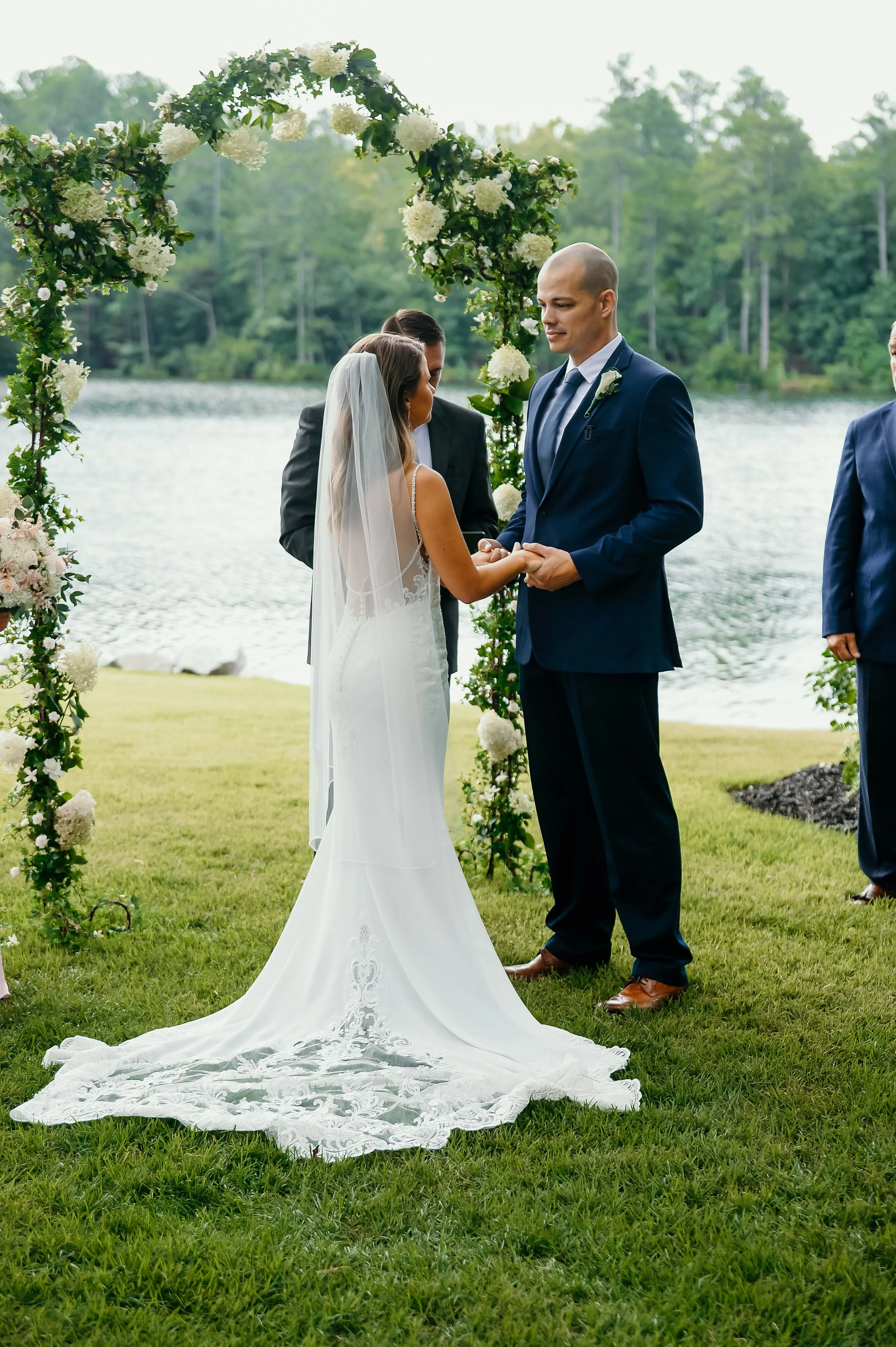A couple getting married outdoors by a lake, with the bride in a white wedding gown and veil, and the groom in a navy blue suit, holding hands under a flower arch canopy.