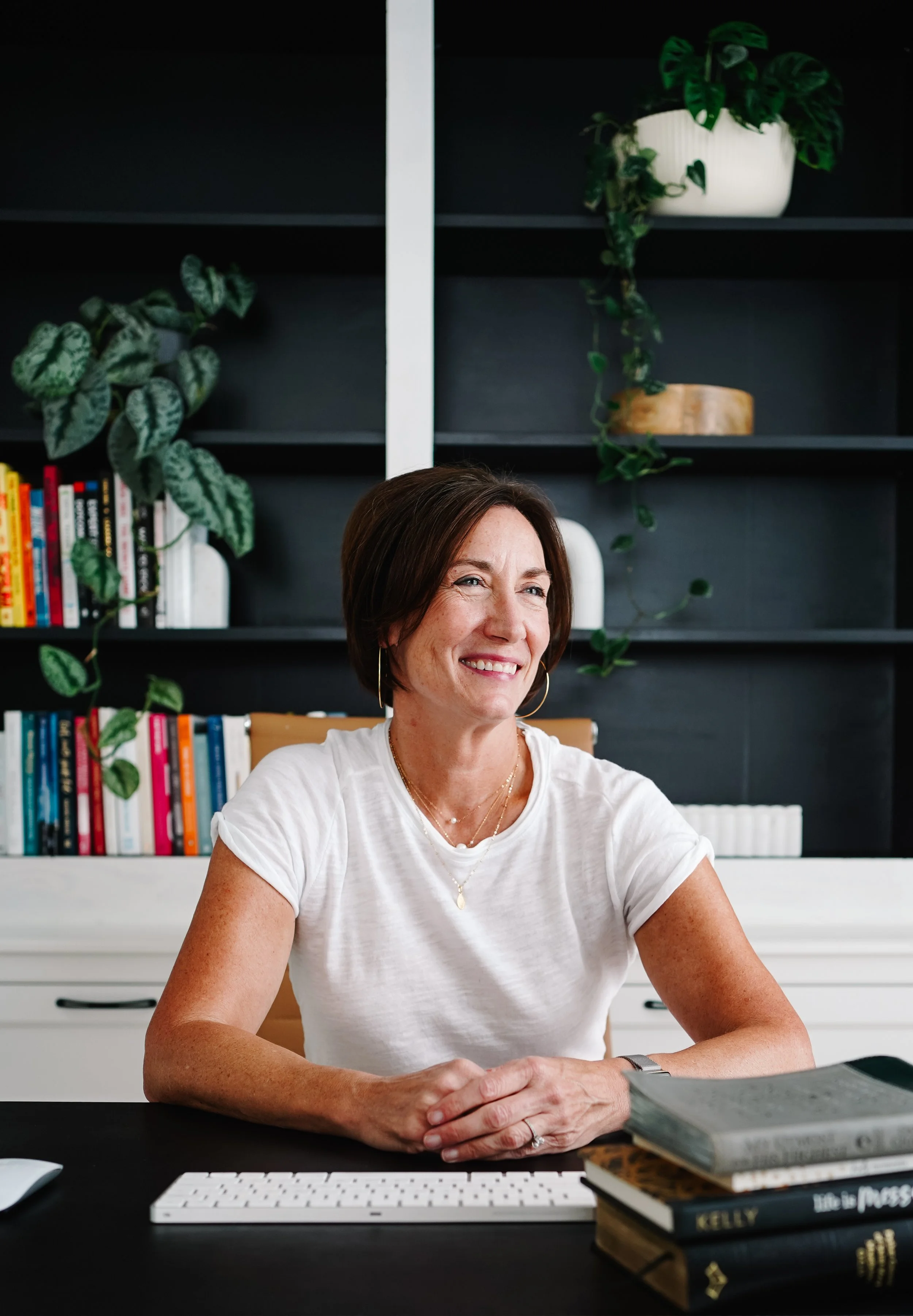 A smiling woman with short brown hair, wearing a white t-shirt, is sitting at a desk with books and a keyboard in front of her, in a room with a dark bookshelf and green plants in the background.