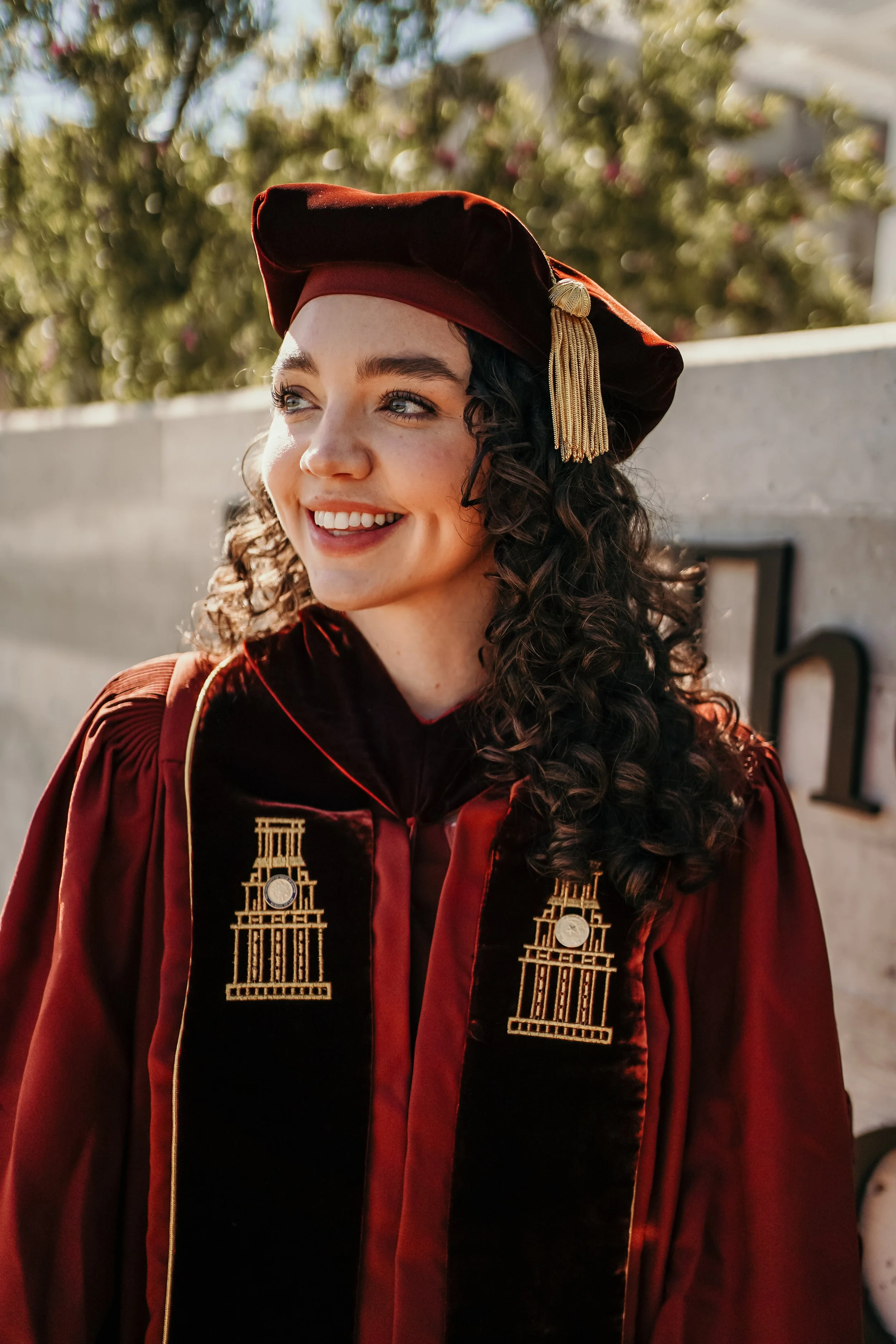 Young woman in a graduation gown and cap smiling outdoors.