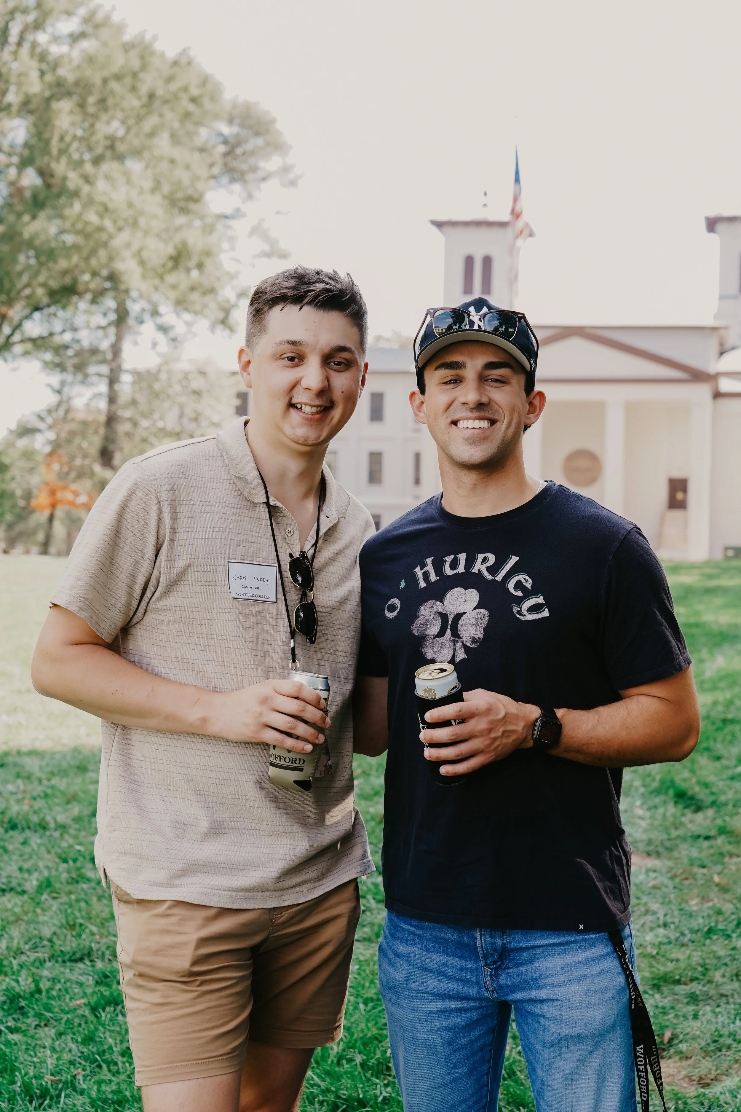 Two young men standing outdoors, smiling, holding drinks, with a historic building and trees in the background.