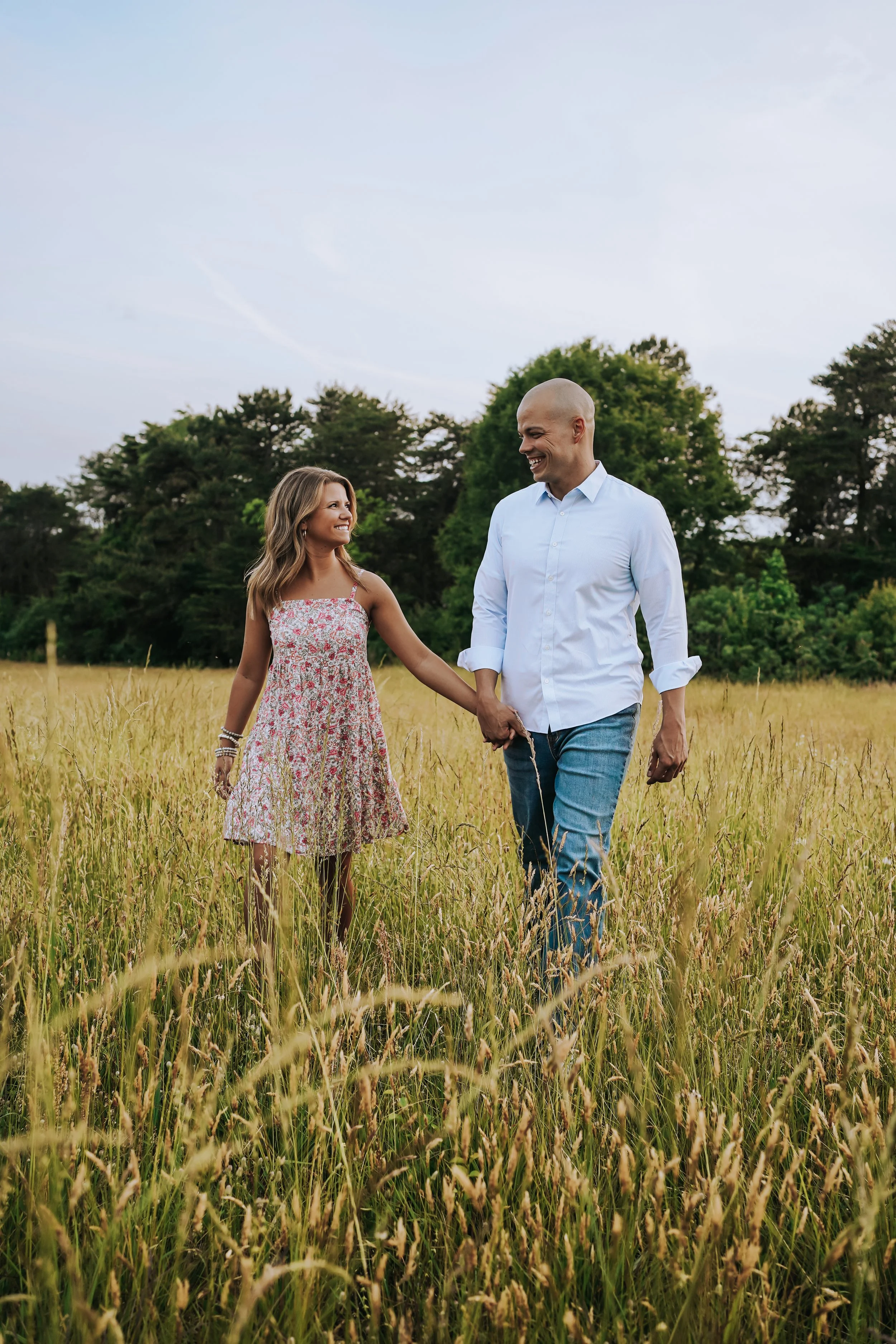 A couple walking hand-in-hand through a grassy field, smiling at each other, with trees and a cloudy sky in the background.