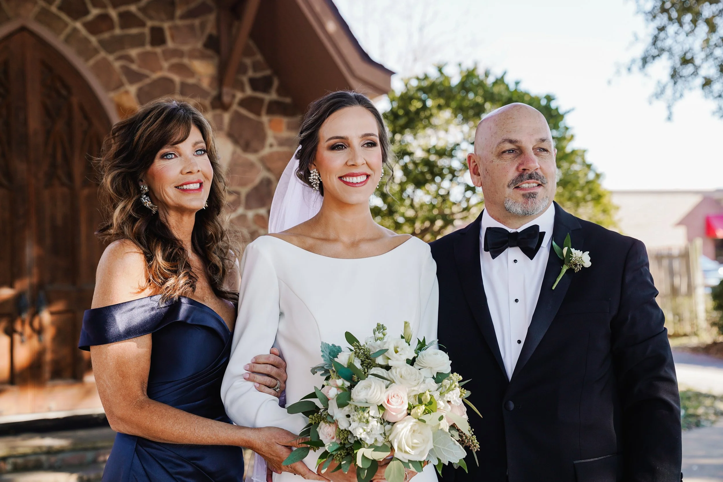 A bride in a white wedding dress holding a bouquet of flowers, smiling, standing between a woman in a navy off-shoulder dress and a man in a tuxedo with a bow tie, outdoors with trees and a stone building in the background.