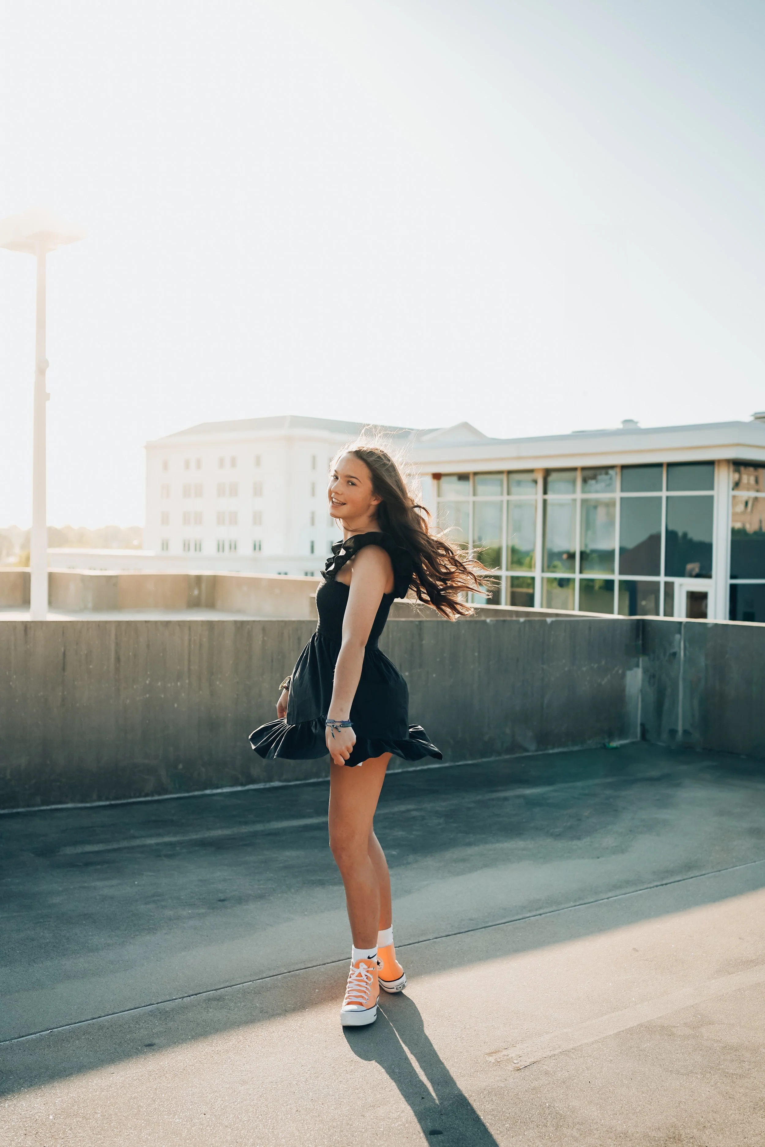 A young woman in a black dress and orange sneakers is standing on a rooftop parking lot, smiling and looking over her shoulder, with buildings and sunlight in the background.