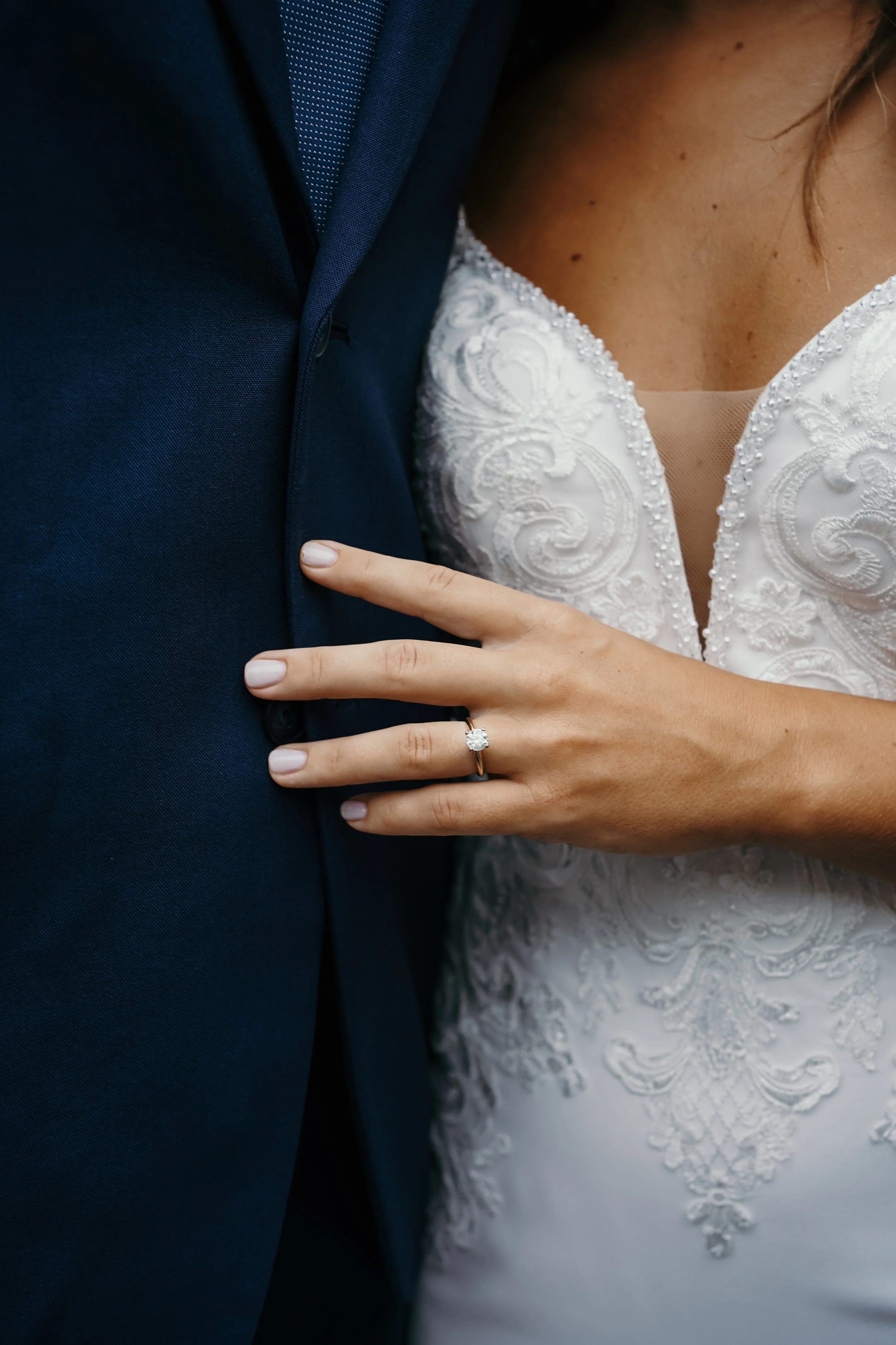 Close-up of a bride's left hand with a diamond ring, resting on a groom's chest, who is dressed in a dark suit. The bride is wearing a white lace wedding dress.