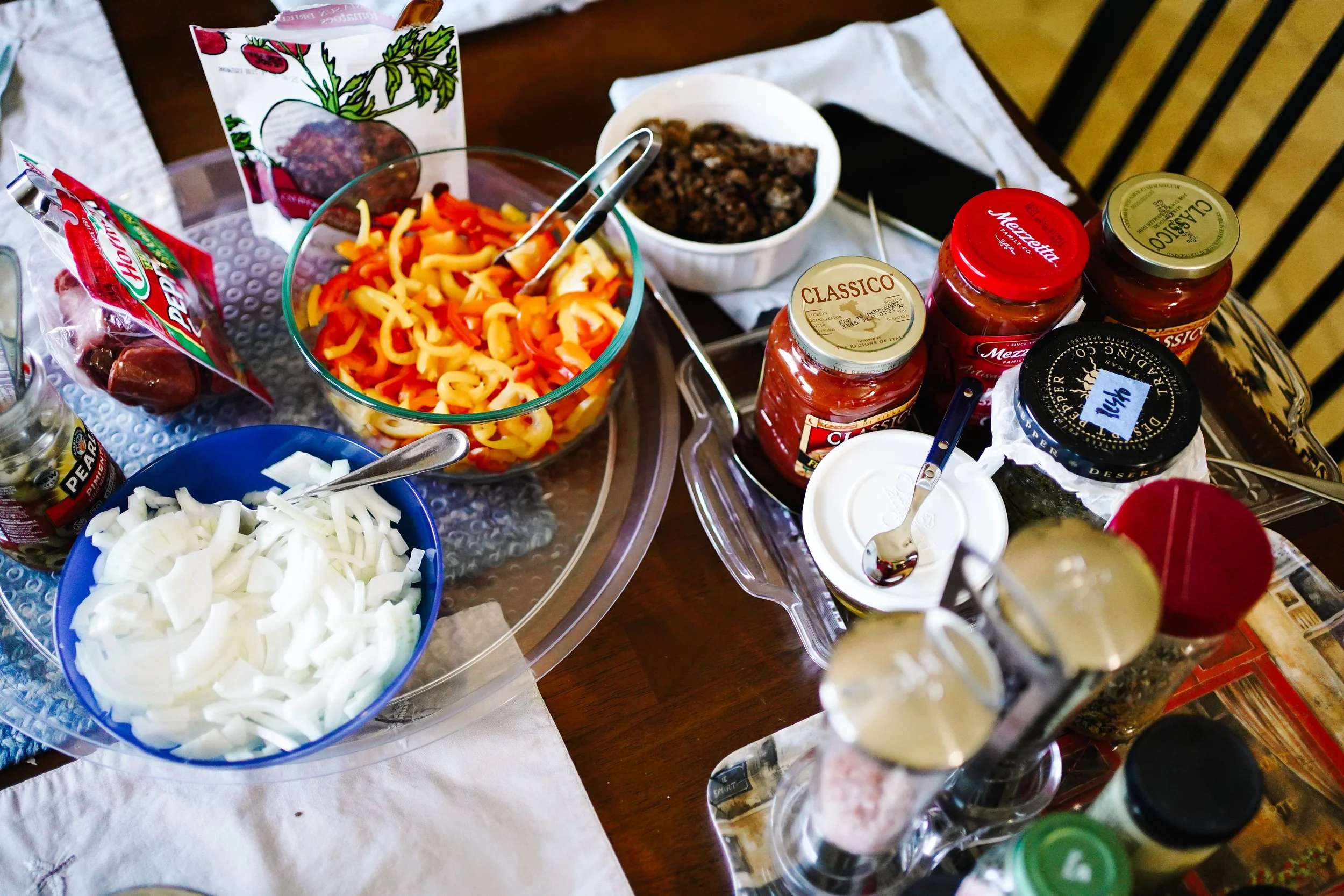 A table set with various ingredients including chopped onions, shredded cheese, sliced bell peppers, jars of pasta sauce, and condiments, preparing for a meal. There are also small bowls and utensils on the table.
