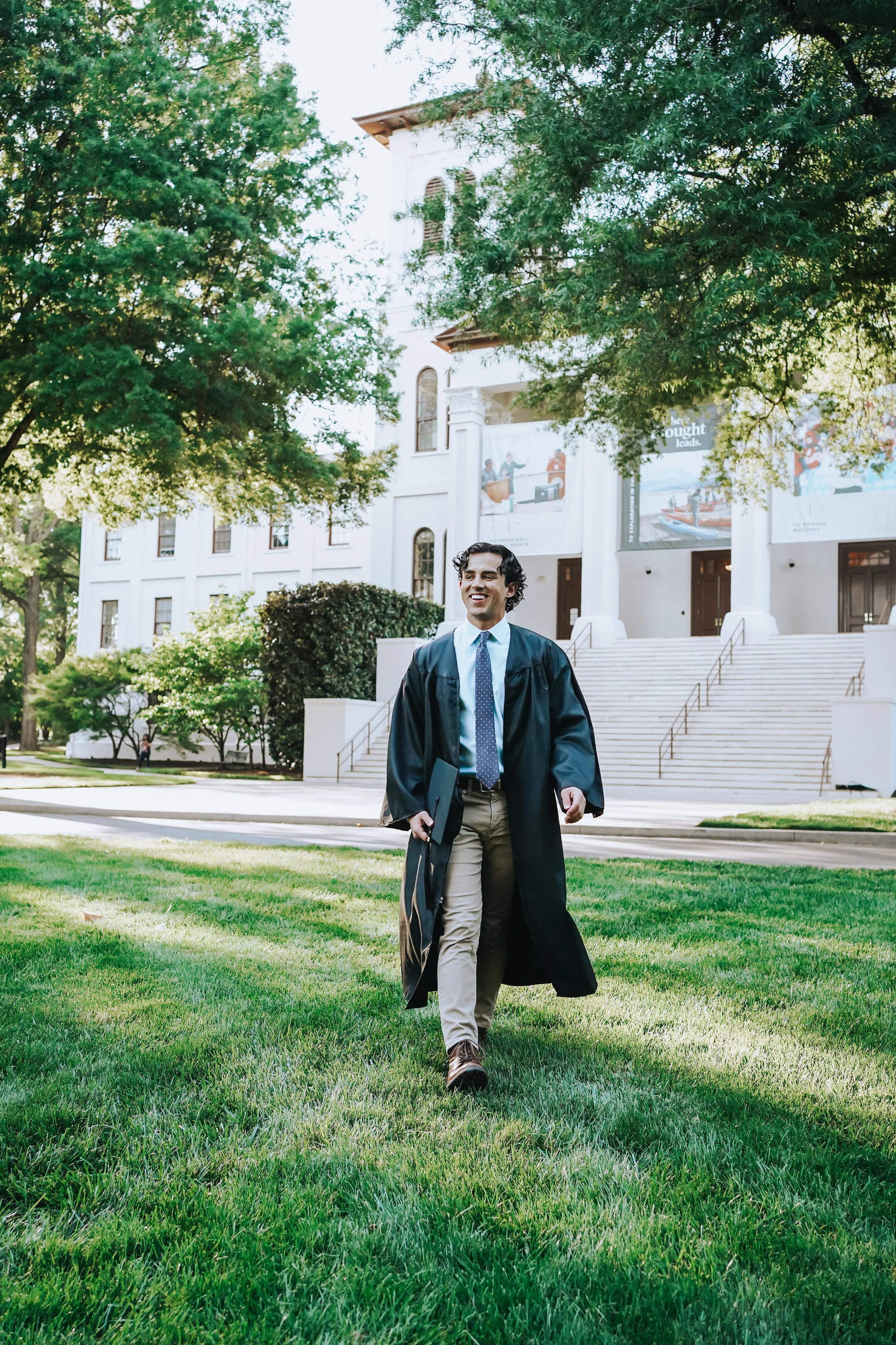 A young man in graduation cap and gown walking on a university campus green, smiling, holding a diploma, with a white institutional building with steps and banners in the background.