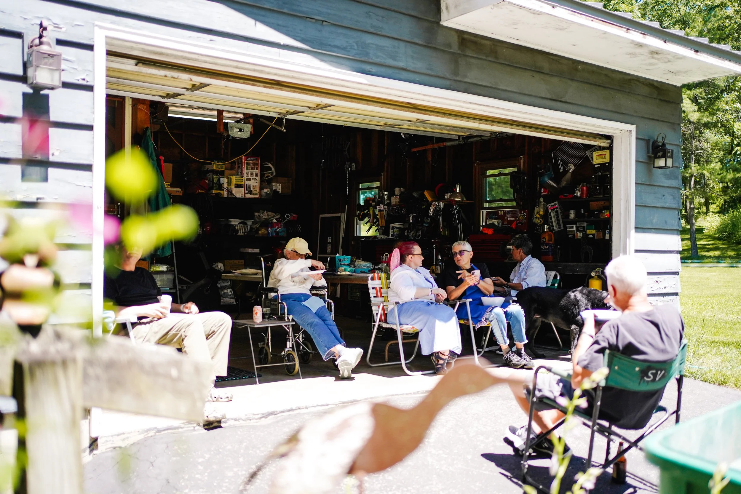 People sitting outside a garage with tools and equipment inside, enjoying a social gathering on a sunny day.