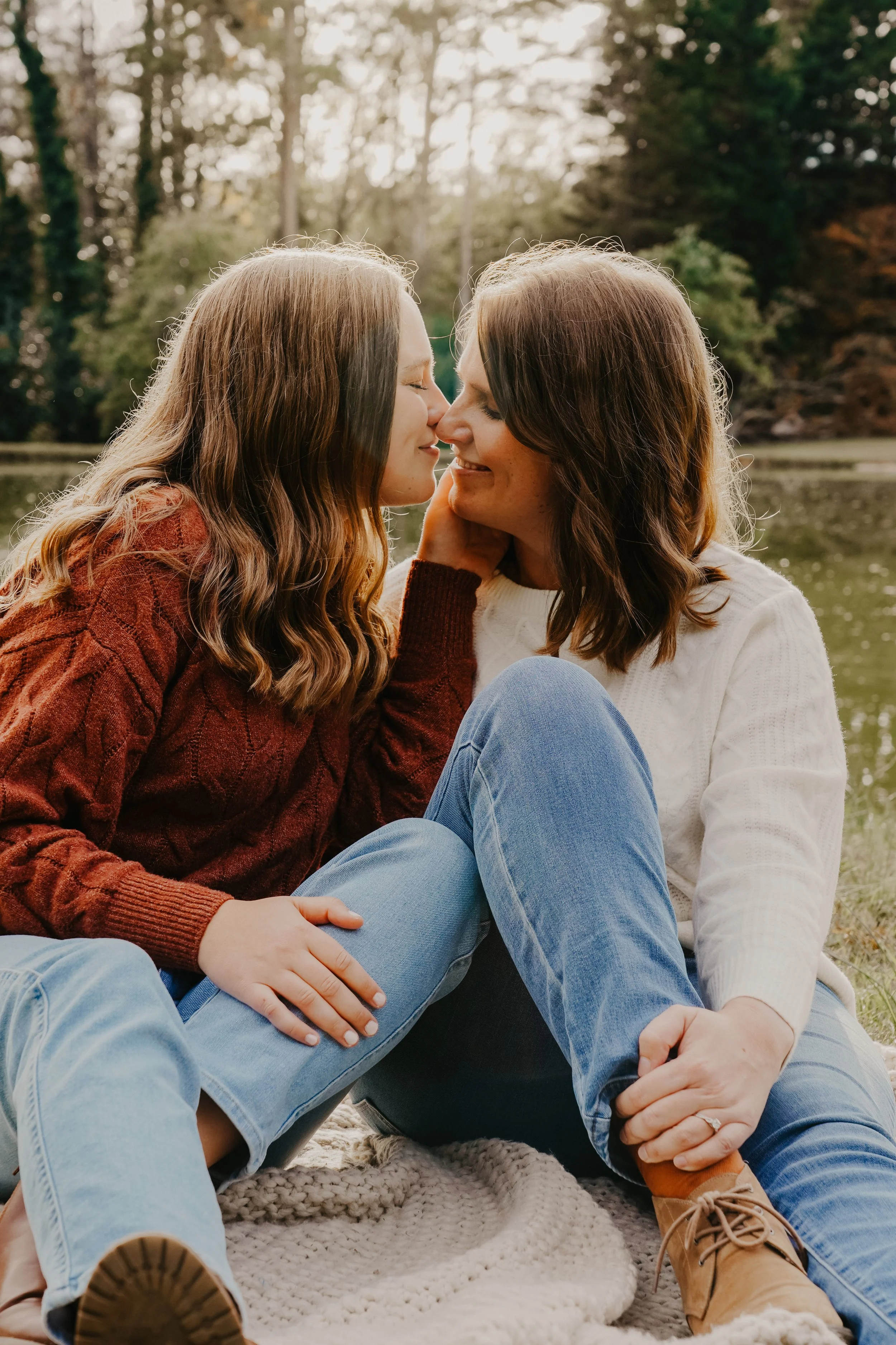 Two women sitting outdoors by a lake, touching noses affectionately and smiling.
