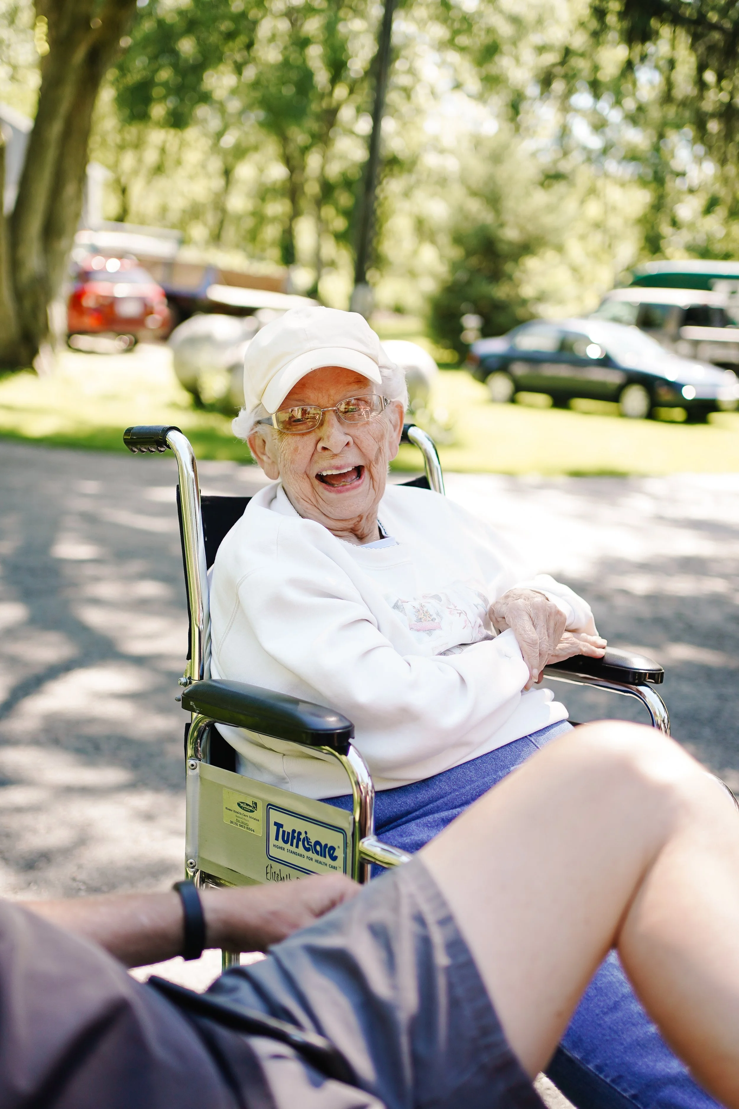 An elderly woman in a wheelchair outdoors, smiling and laughing, with trees and parked cars in the background.