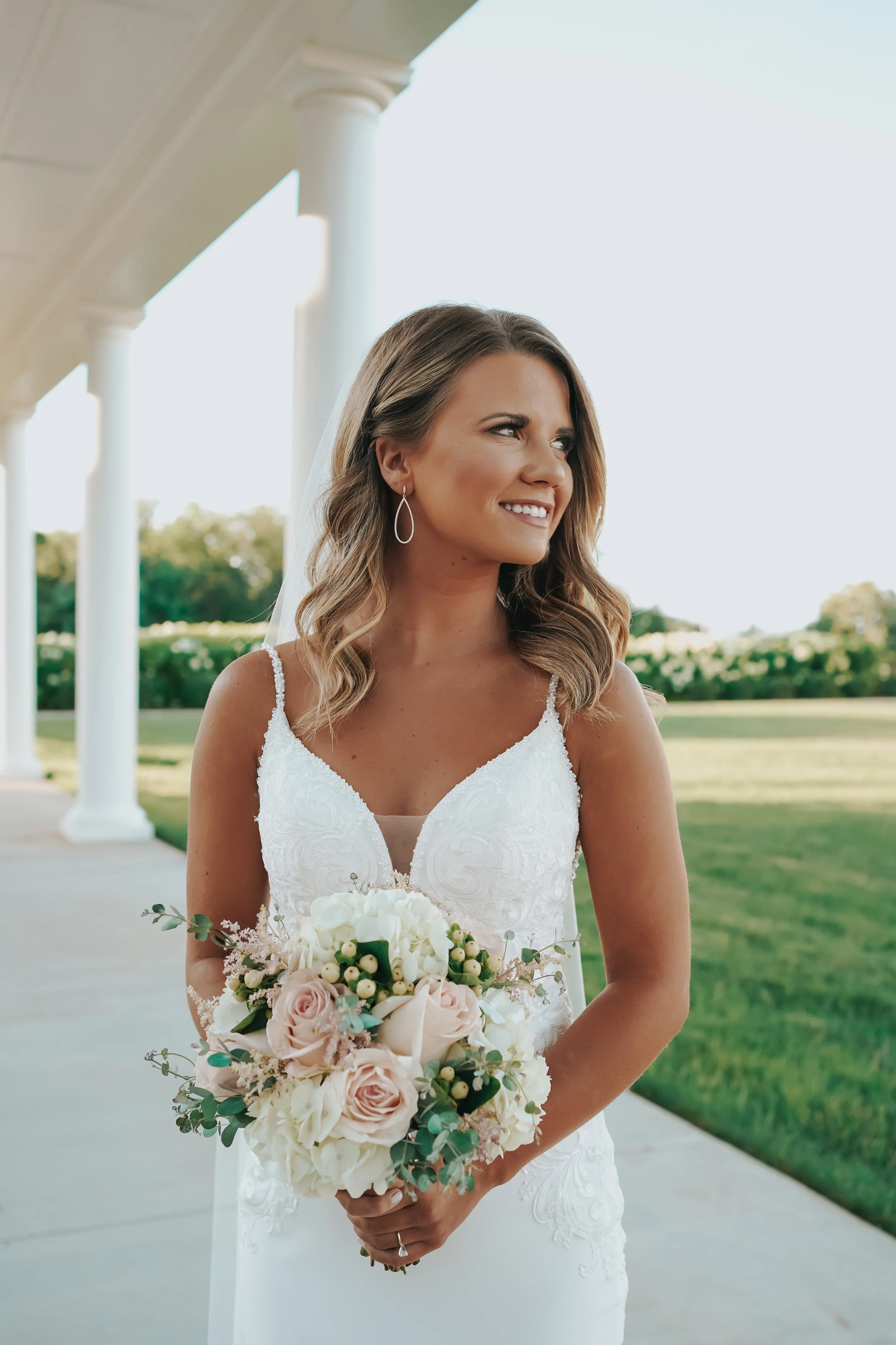 A bride in a white wedding dress holding a bouquet of pink and white roses outdoors on a sunny day.