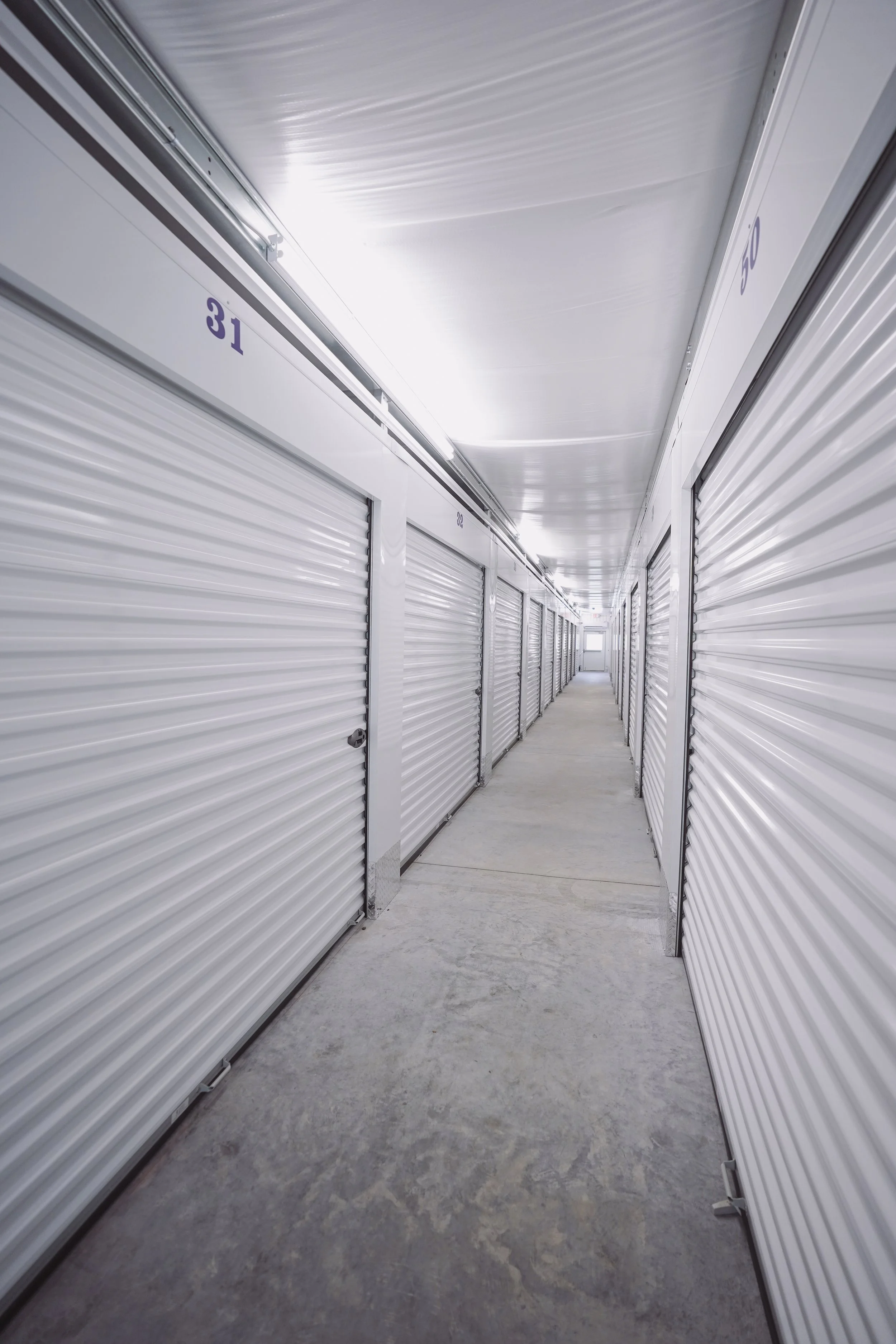 Long corridor of storage unit lockers with white metal doors and numbered labels, illuminated by fluorescent lighting.