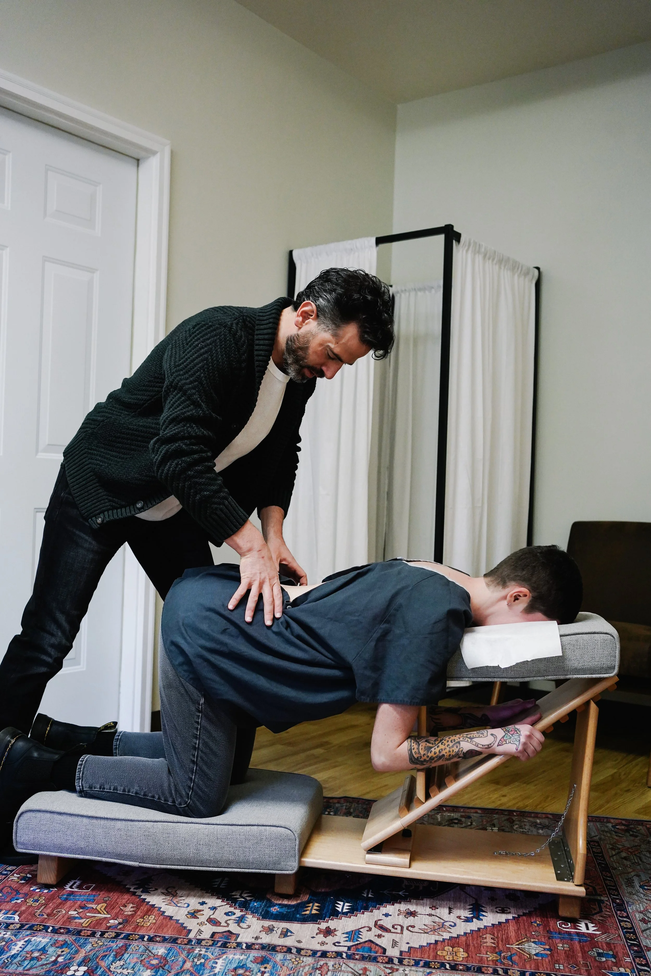A man is giving a chiropractic adjustment to a young man who is kneeling on a padded bench with his head resting on a cushioned support, in a room with a wooden floor, a rug, and a privacy curtain.