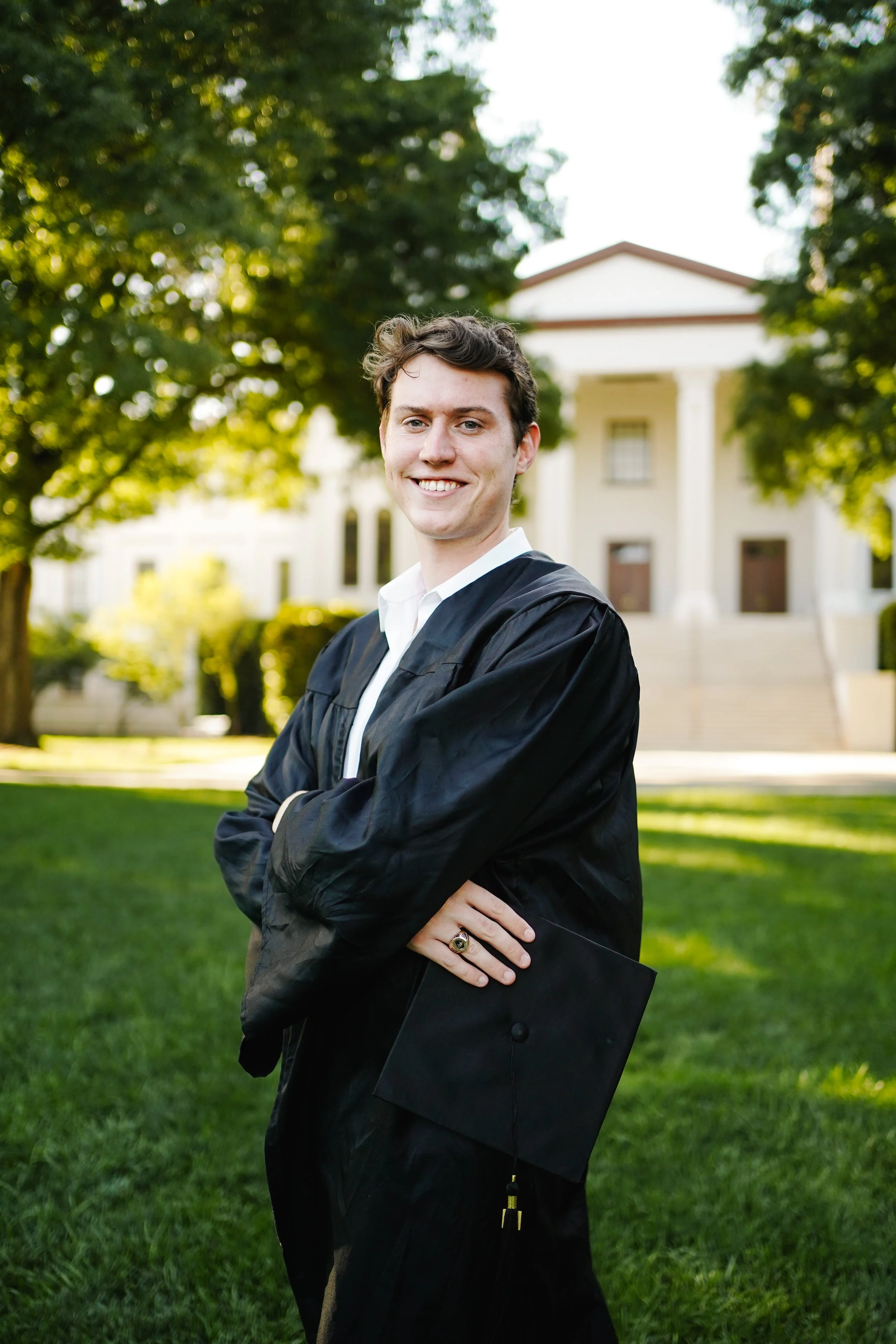 A young man in a black graduation gown standing outdoors on a grassy area with a large white building and trees in the background.