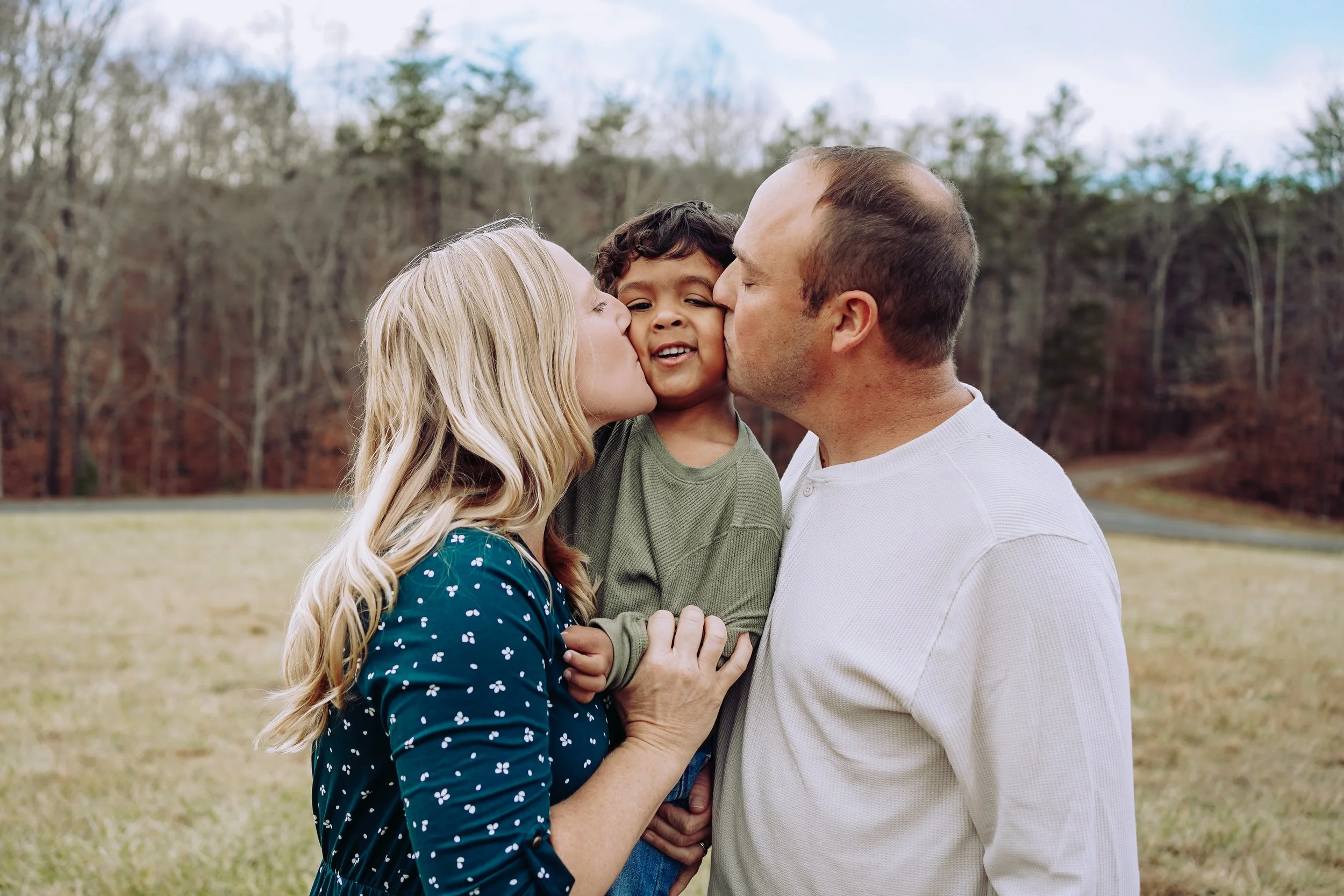 A family of three, a mother, father, and young son, sharing kisses outdoors in a grassy field with trees in the background.