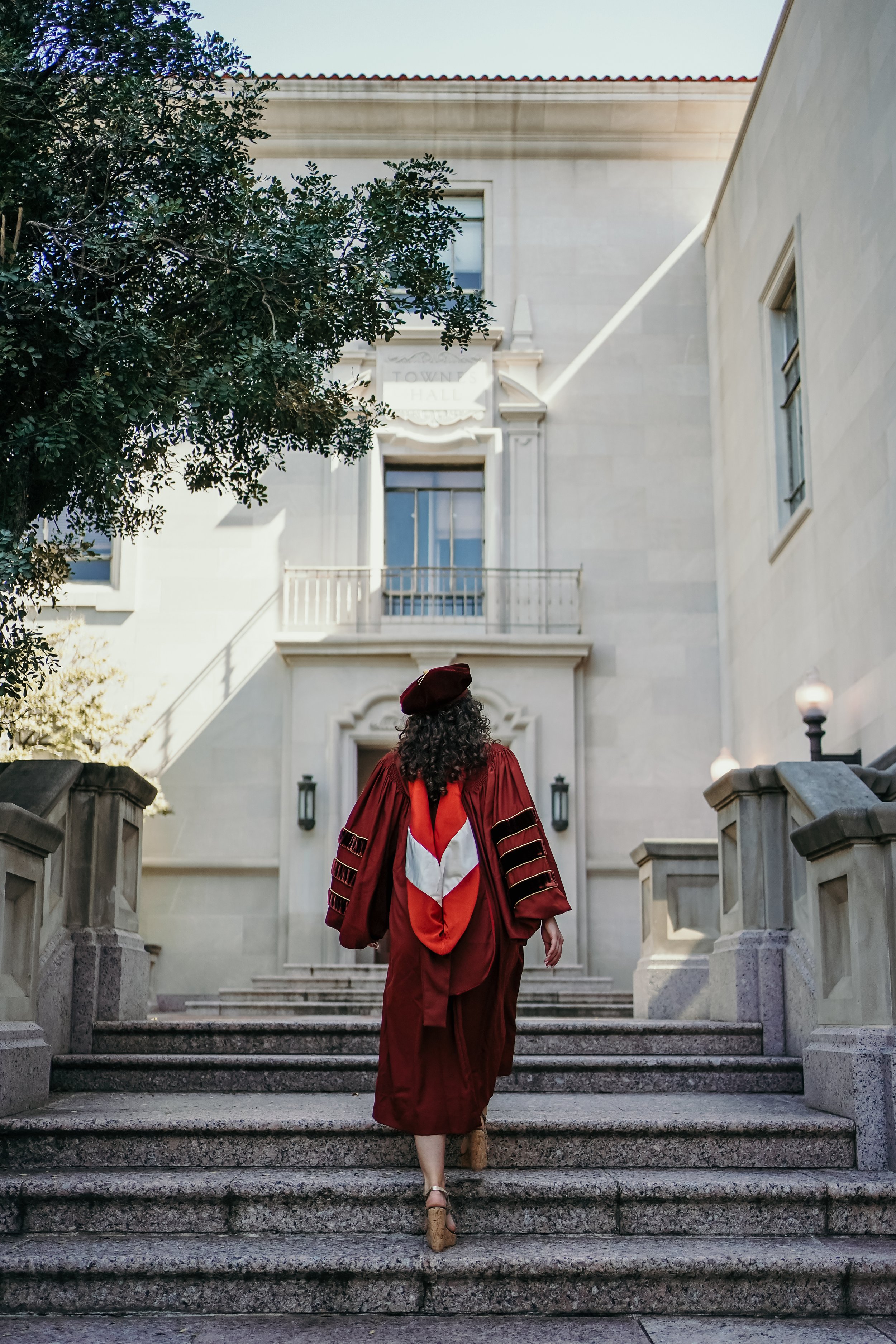 Woman in academic regalia climbing stairs towards building, likely at graduation ceremony.