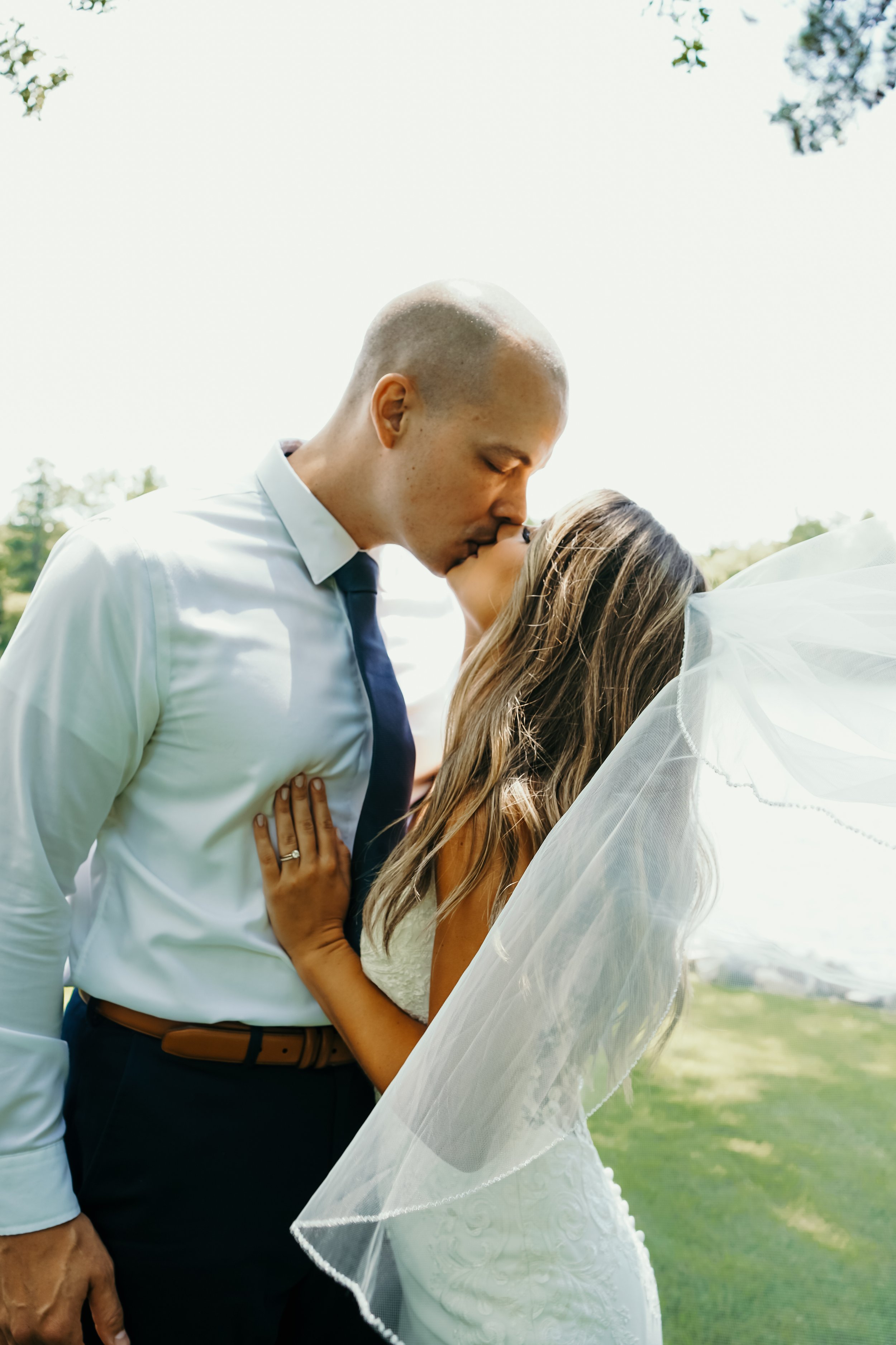 A newlywed couple sharing a kiss outdoors, with the groom wearing a white shirt and navy tie, and the bride in a white wedding dress and veil, embracing against a green landscape.