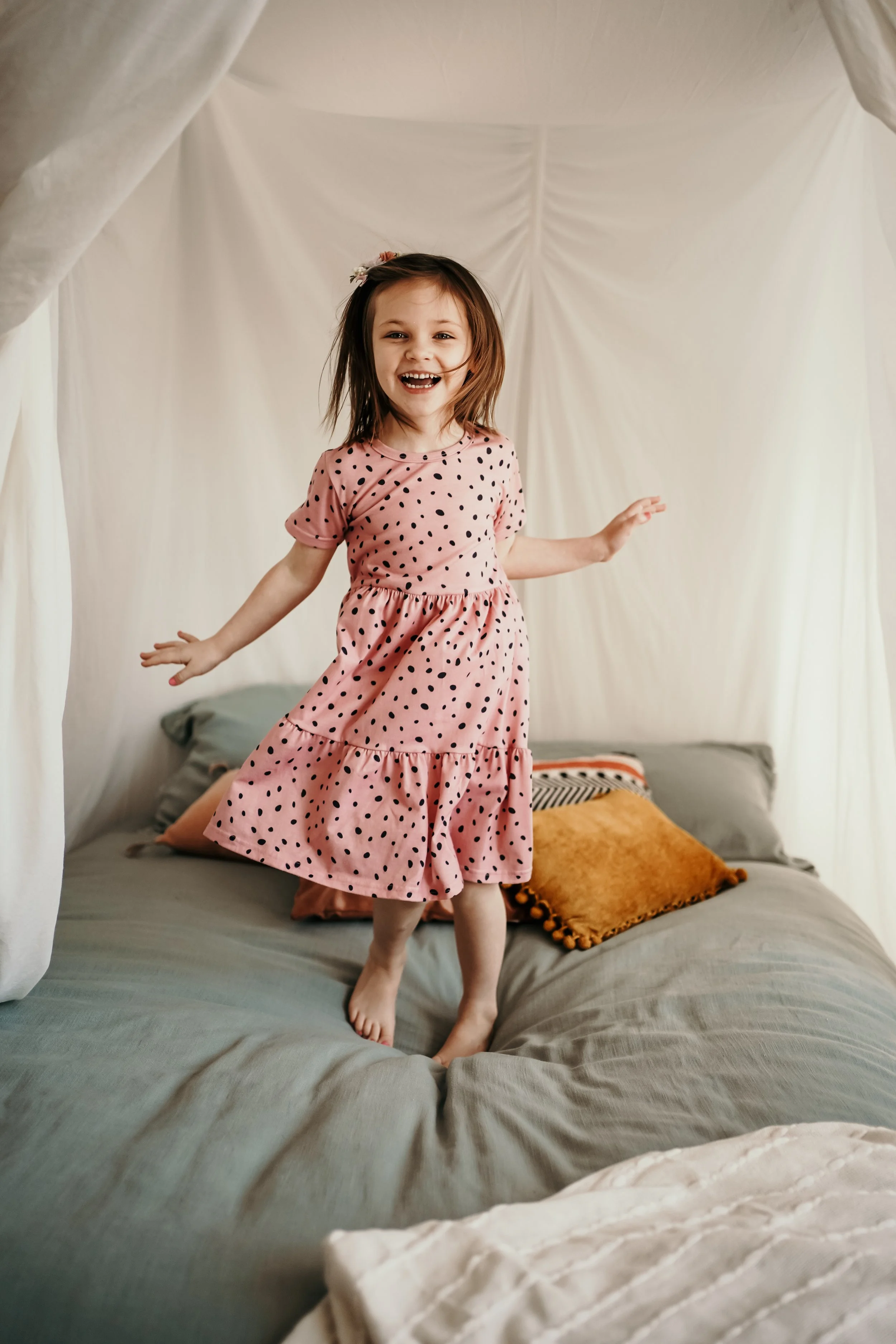 A young girl with brown hair, wearing a pink polka dot dress, jumping on a bed with pillows and a white canopy in the background.