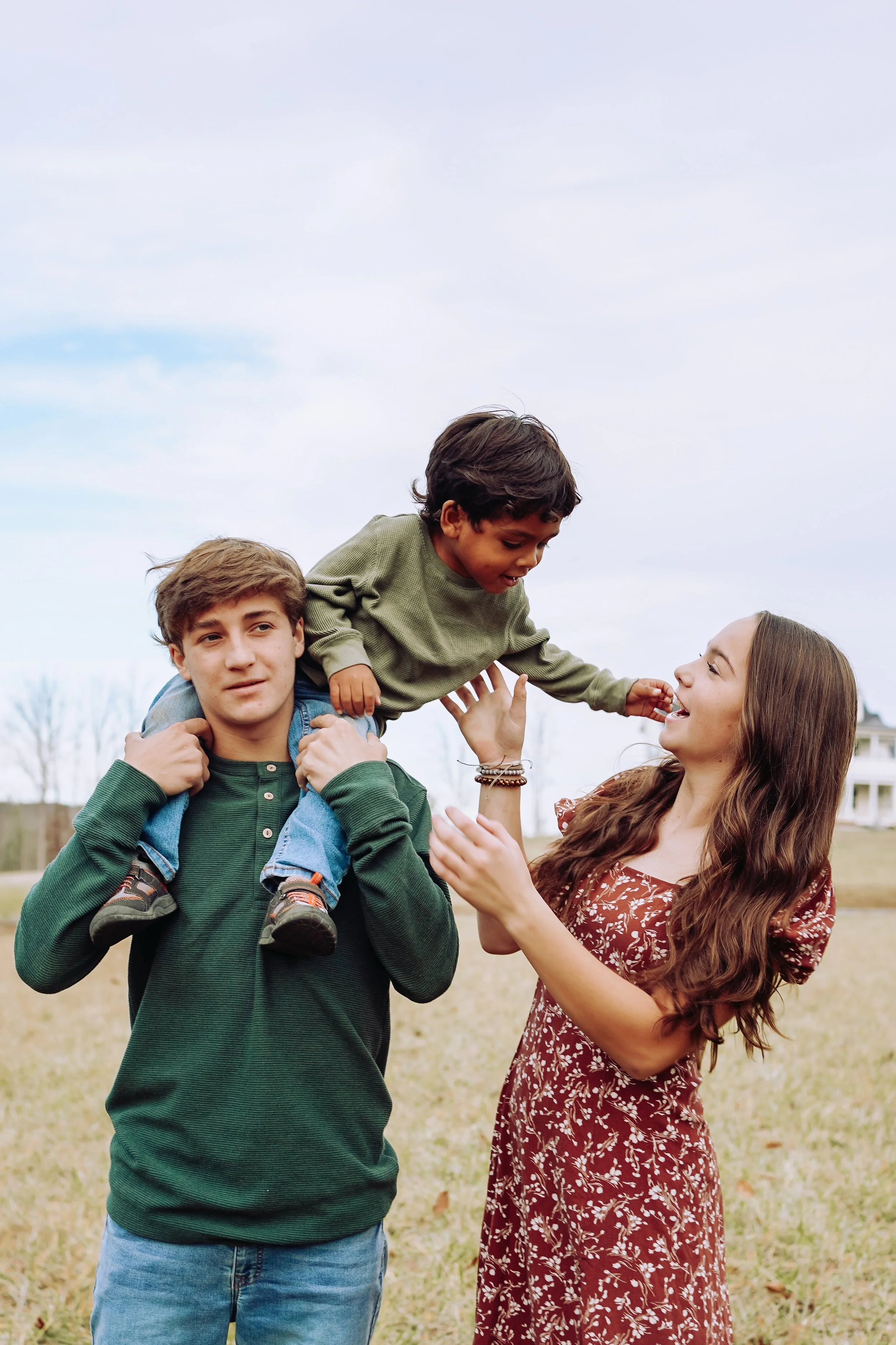 A woman with long brown hair in a floral dress holding a young boy with brown hair on her hand while a teenage boy with brown hair in a green hoodie carries a toddler boy on his shoulders, all outdoors on a grassy area on a cloudy day.