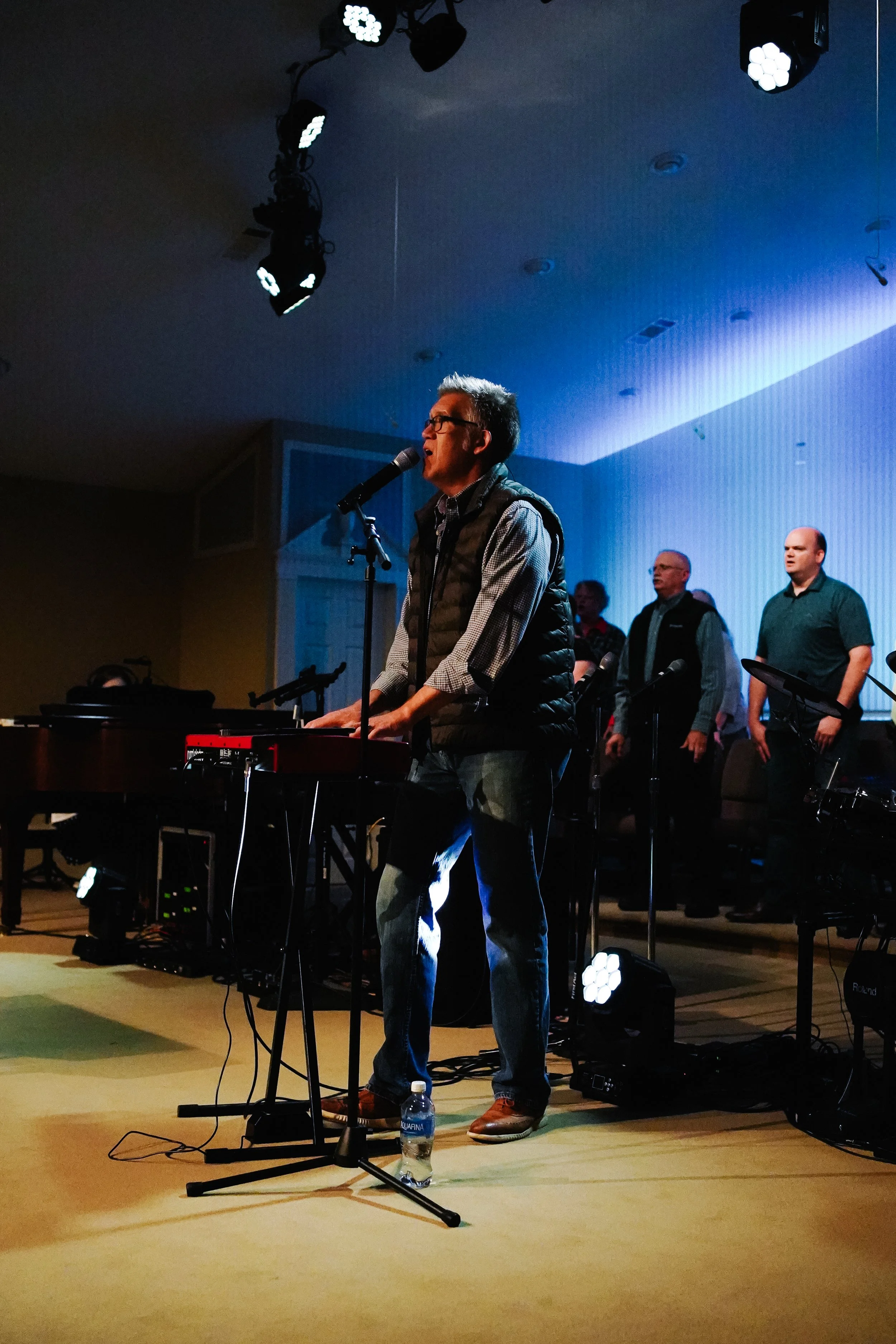 A man singing and playing keyboard on stage with four men standing behind him, stage lighting overhead, and a water bottle on the floor.